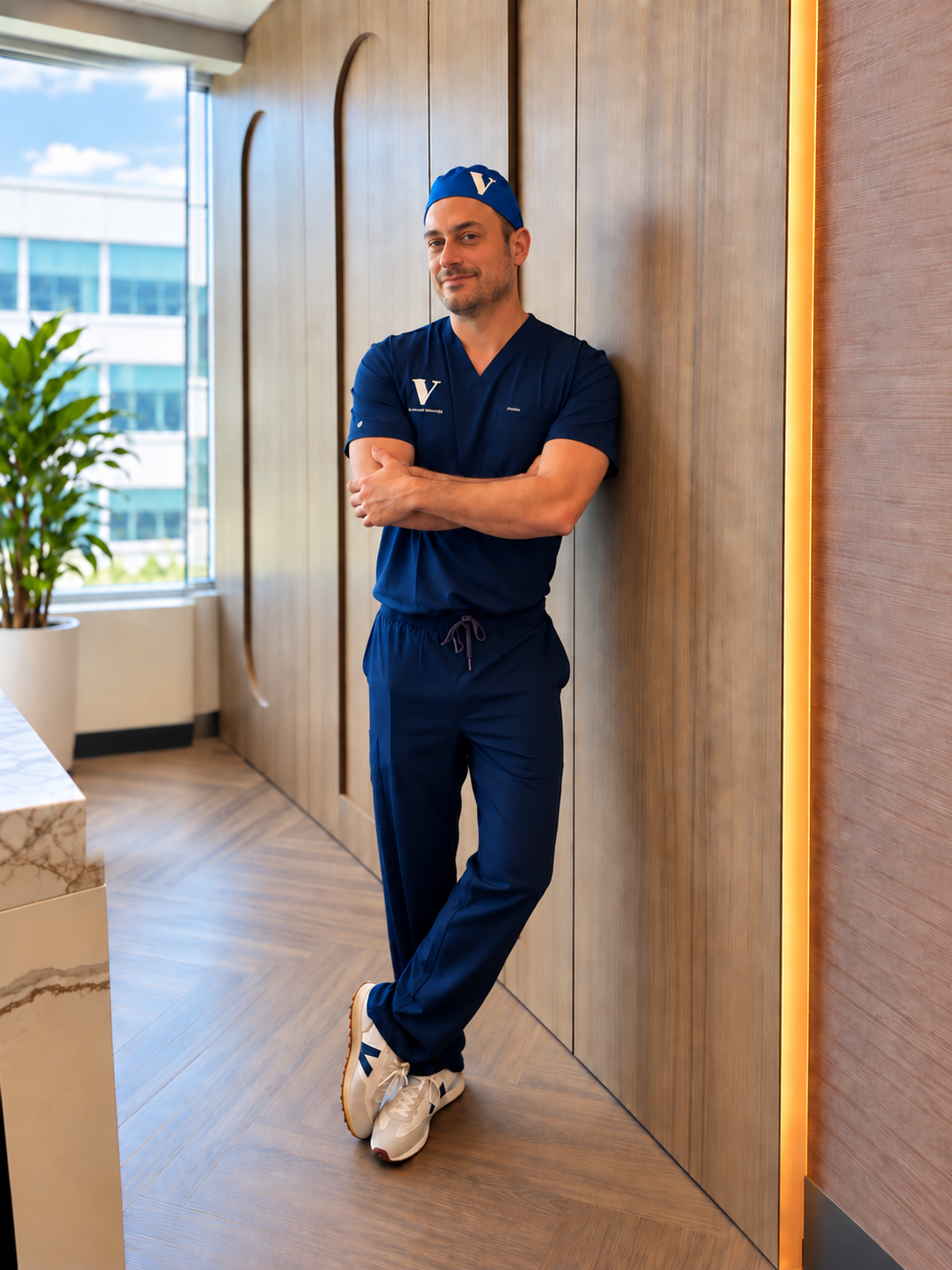 Dr Michael Secter - A man wearing a navy blue medical uniform and cap, with a 'V' logo, leaning against a wooden wall in a modern office space with a large window and a green plant.