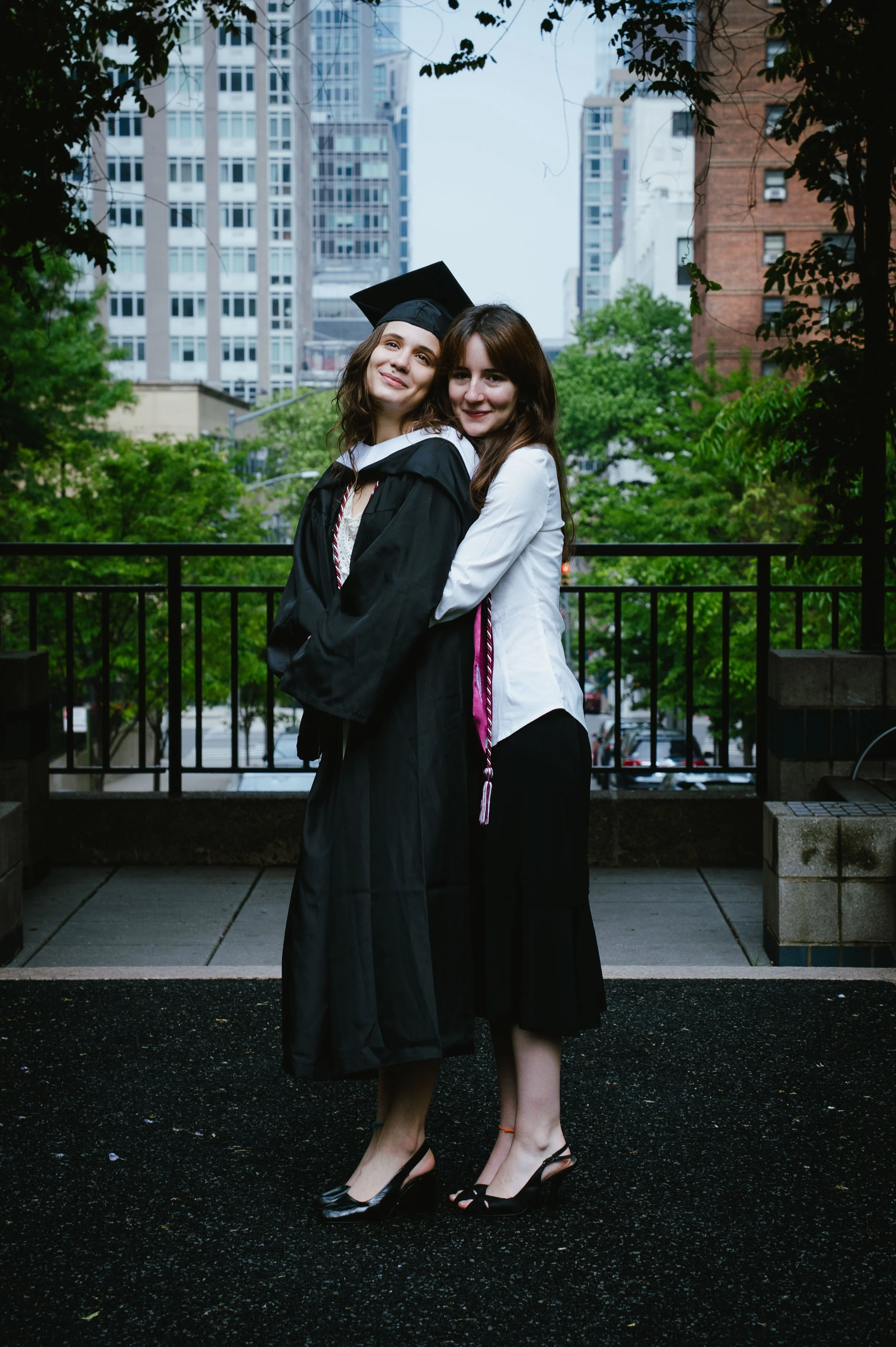Two young women standing together outdoors in an urban setting, one wearing a graduation cap and gown, and the other in formal attire. They are embracing each other and smiling at the camera, with tall buildings and green trees in the background.