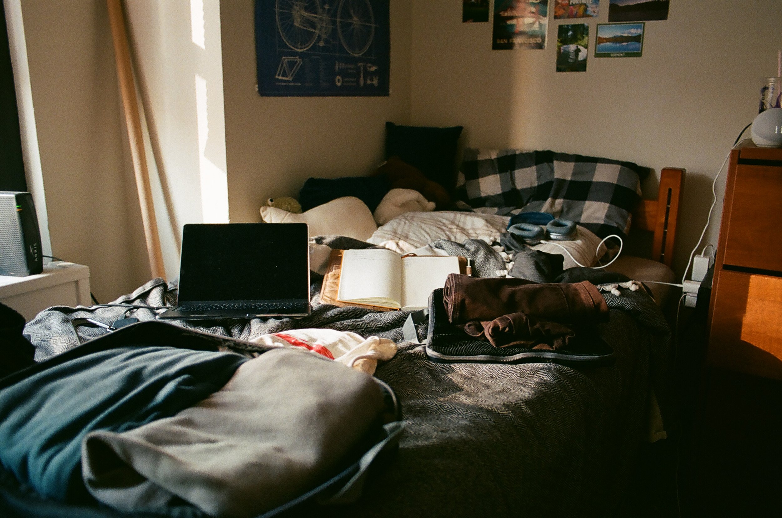 A cluttered bedroom viewed from a person's perspective lying on the bed. The bed has various clothes, an open notebook, a laptop, and a wallet. The wall behind has photos and posters, including a bicycle blueprint. There's a window on the left letting in light, a small white nightstand with a speaker, and a dresser on the right with a smart speaker and a lamp.