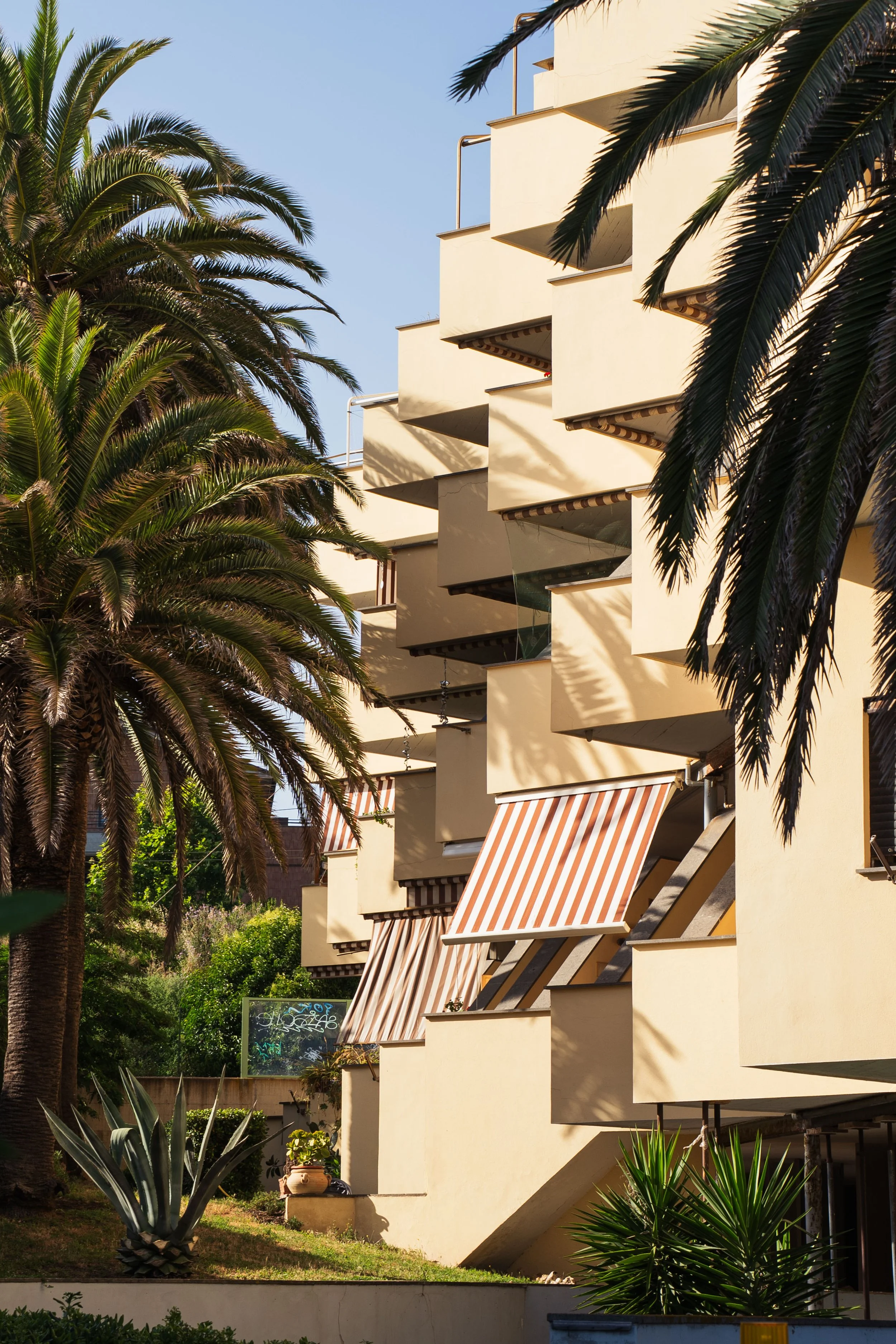 Multi-story beige building with striped awnings on the balconies, surrounded by tall palm trees and various plants, under a clear blue sky.