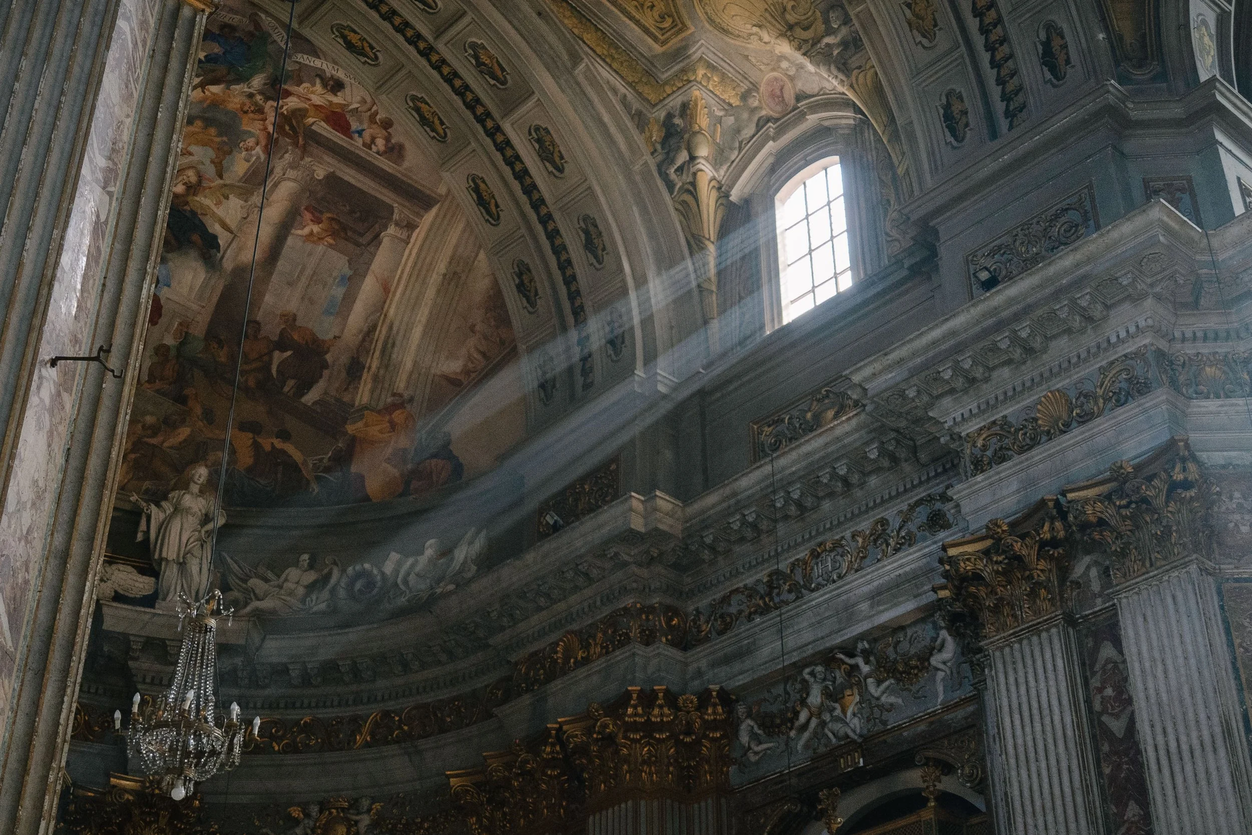 The interior of a grand historic church or cathedral with ornate gold and white decorative elements, a large window allowing sunlight to stream in, and intricate ceiling murals.