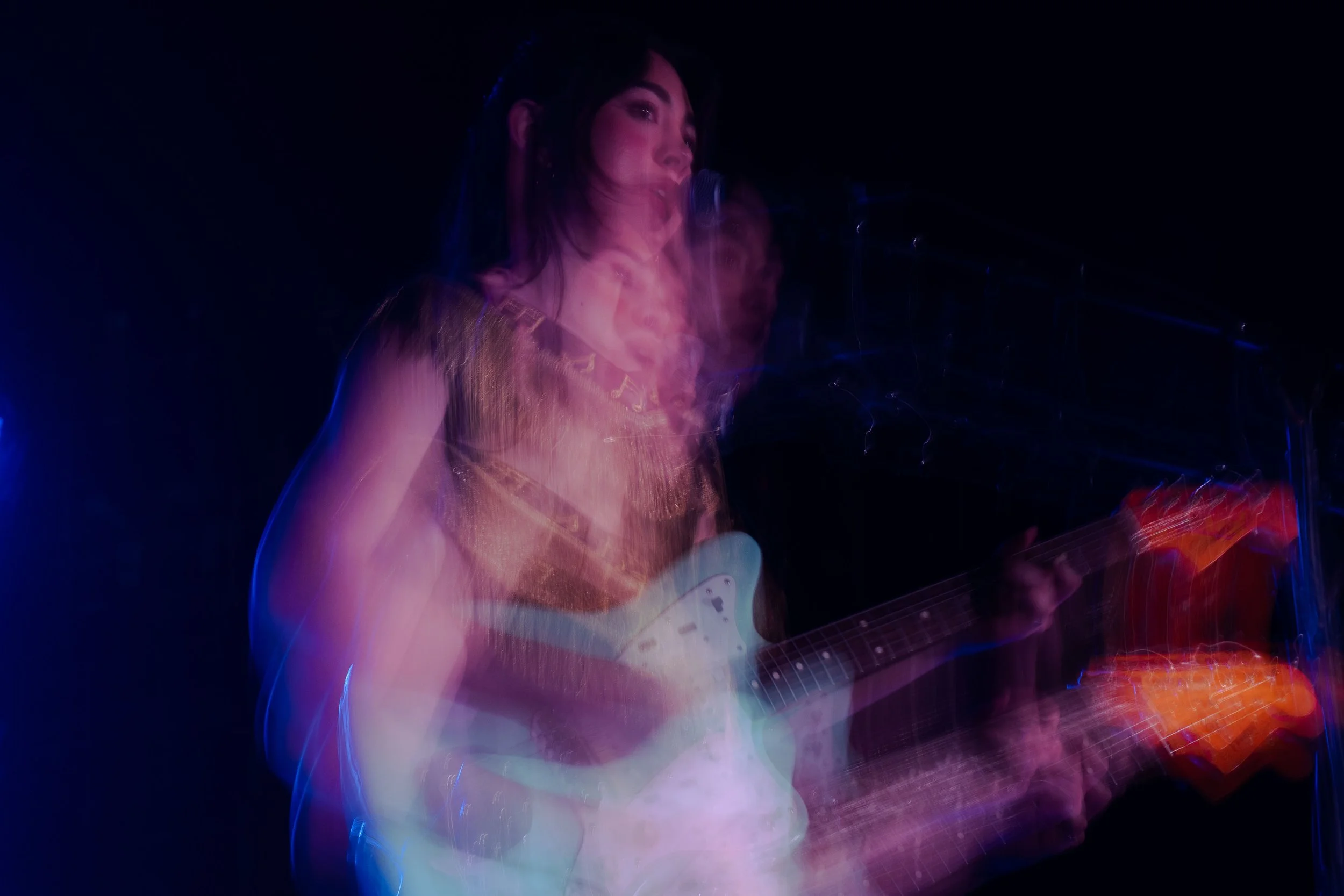A woman singing into a microphone while playing an electric guitar, with colorful light streaks creating a motion effect against a black background.