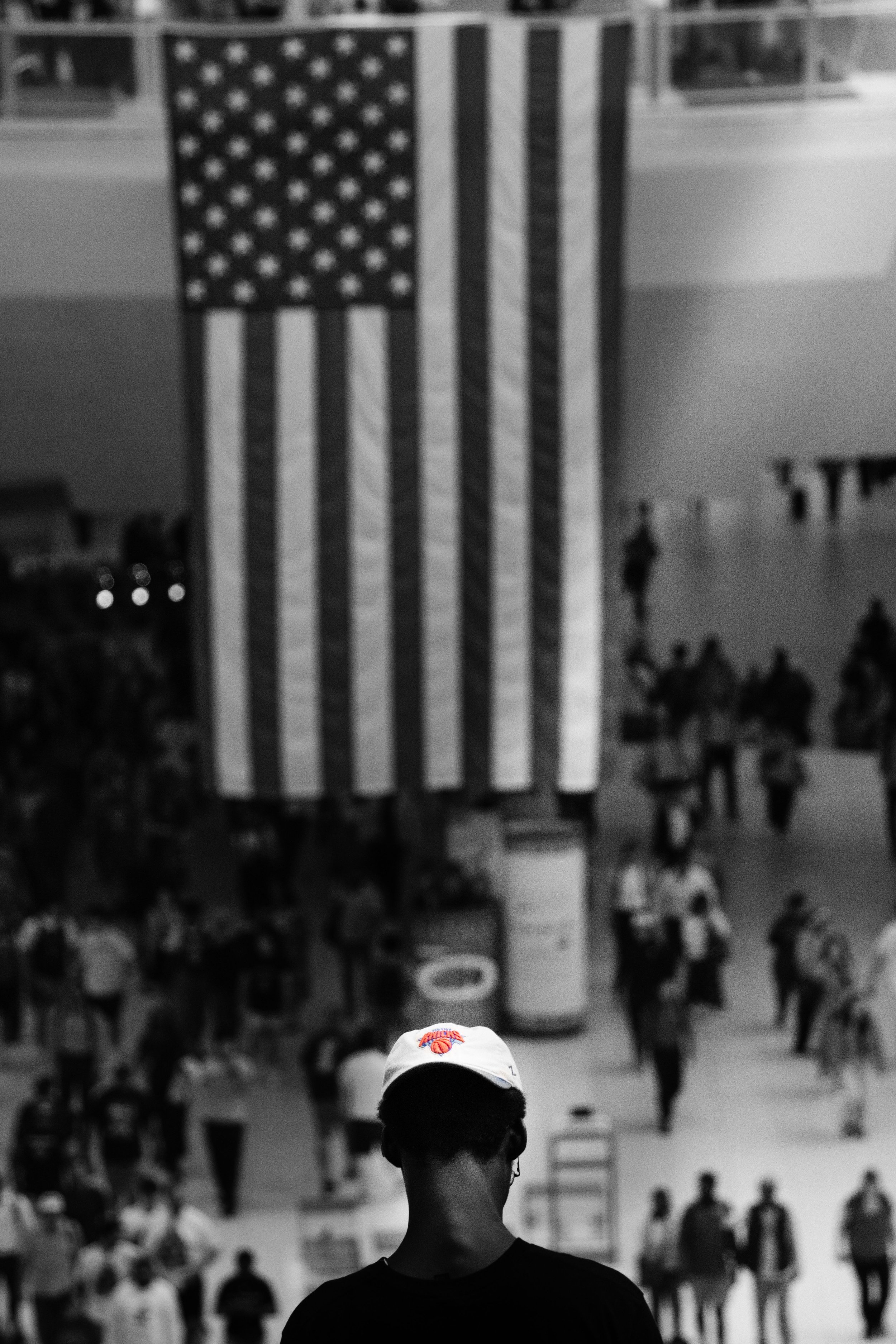 A person wearing a cap viewing a large American flag hanging in a gymnasium filled with people.