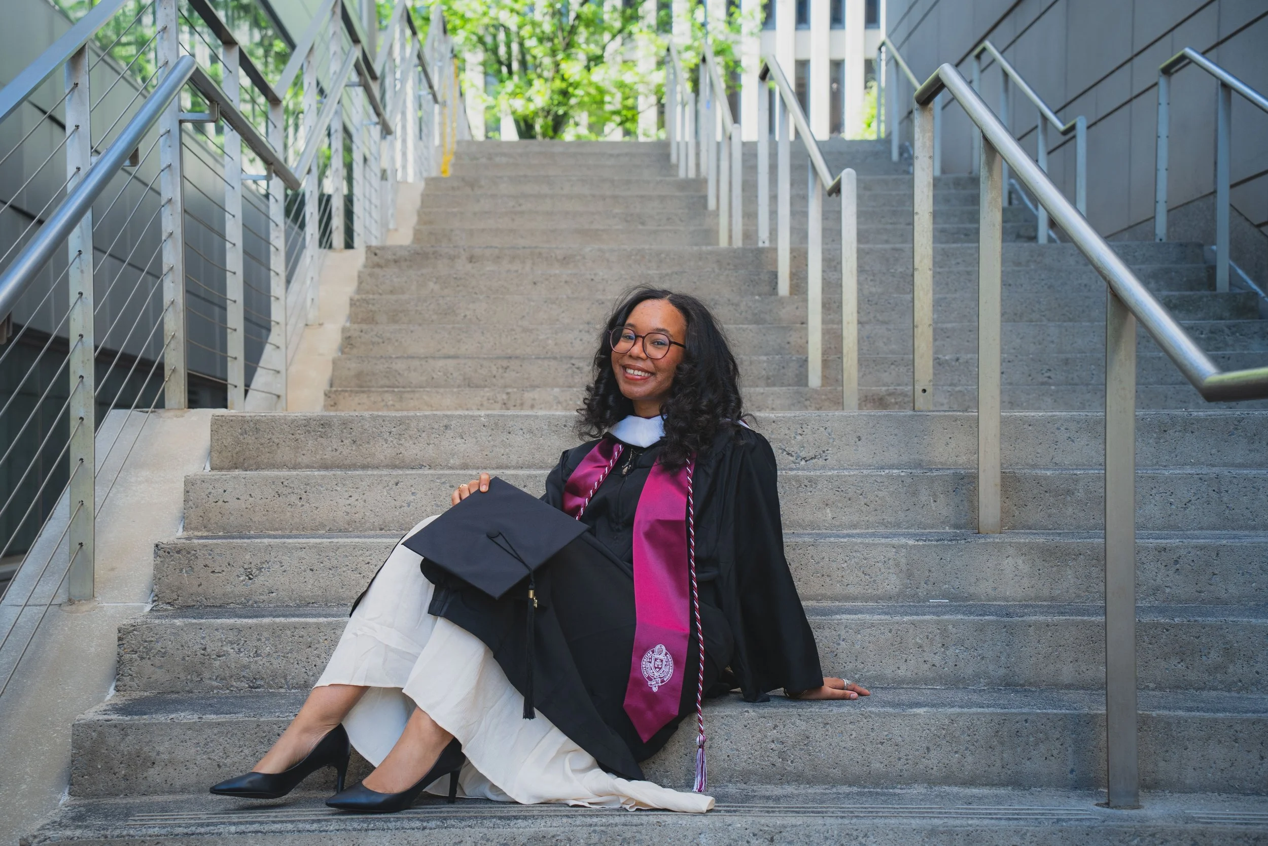 A woman in graduation attire, sitting on outdoor concrete stairs, smiling, with trees in the background.