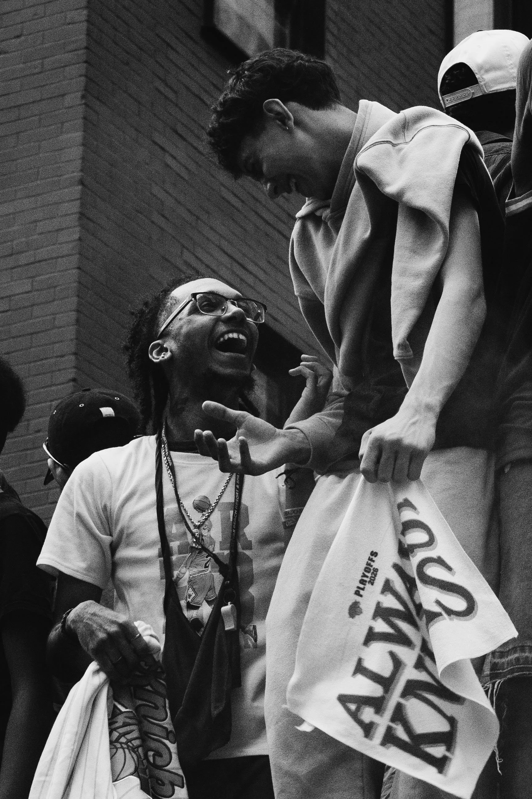 Two young men laughing and engaging with each other. One is wearing glasses, a white t-shirt, and necklaces, while the other is wearing a hoodie and holding a poster or banner.