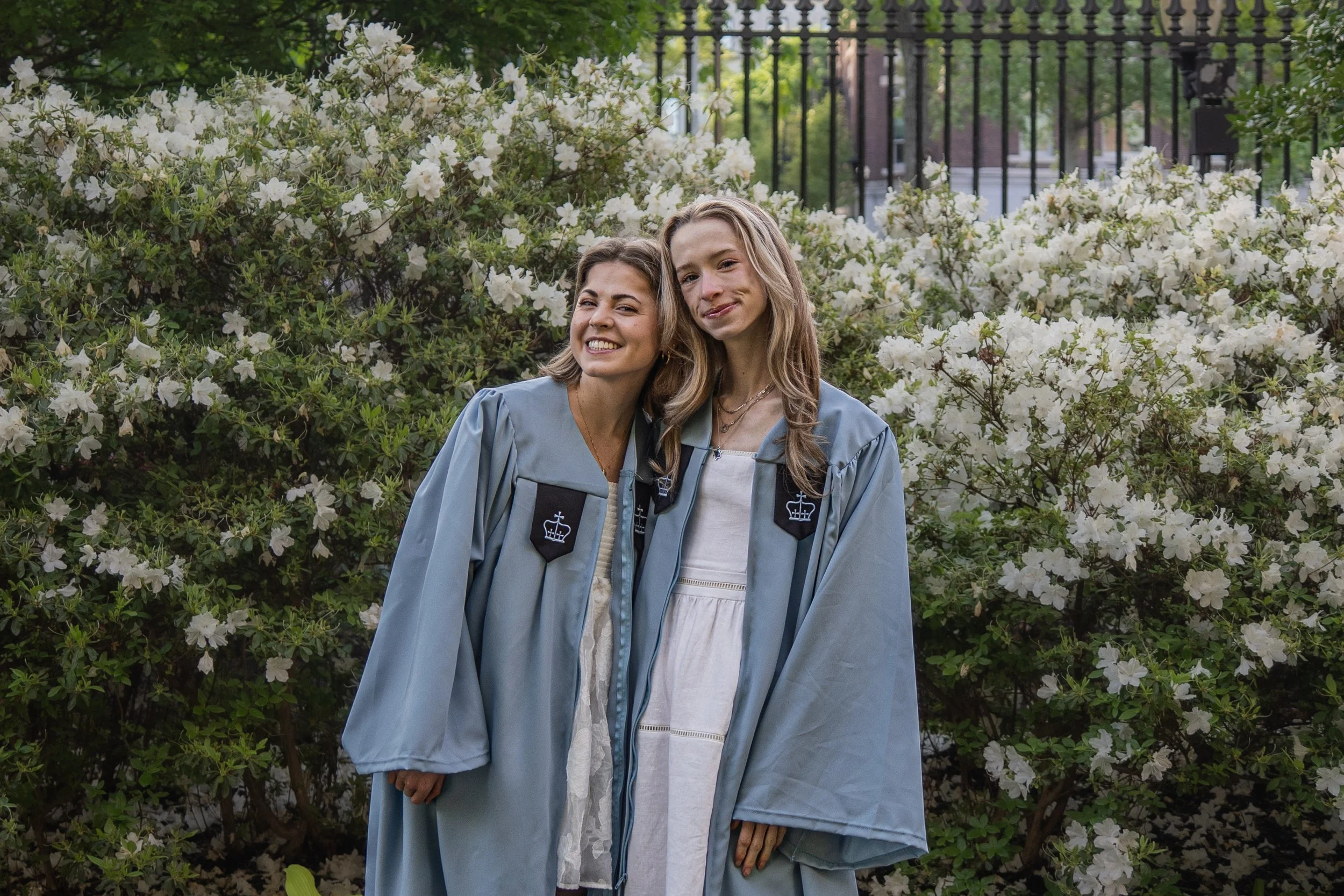 Two young women in graduation gowns standing in front of white flowering bushes, smiling at the camera.