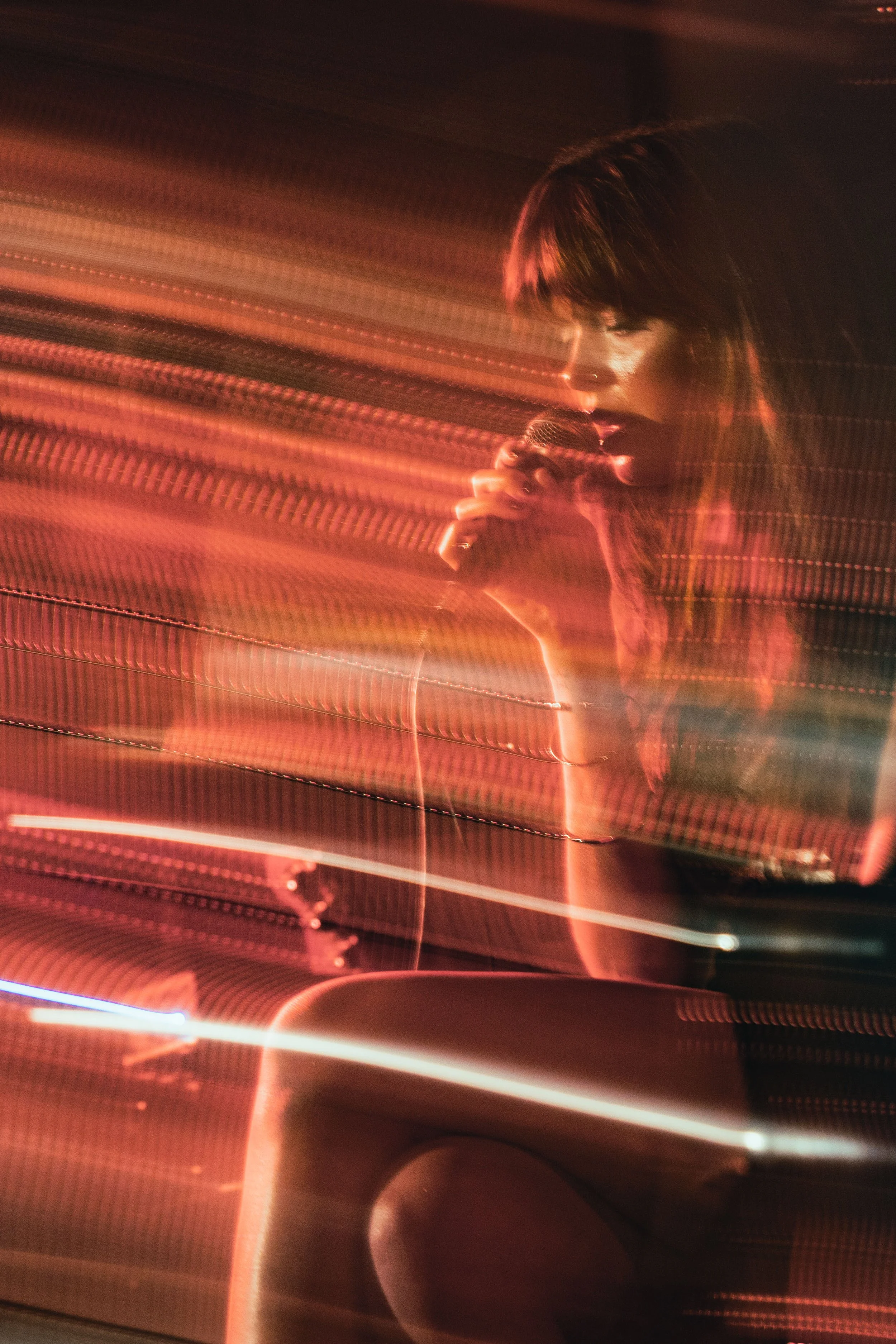 A woman with short, dark hair performing on stage, holding a microphone, with colorful light trails and reflections creating a dynamic background.