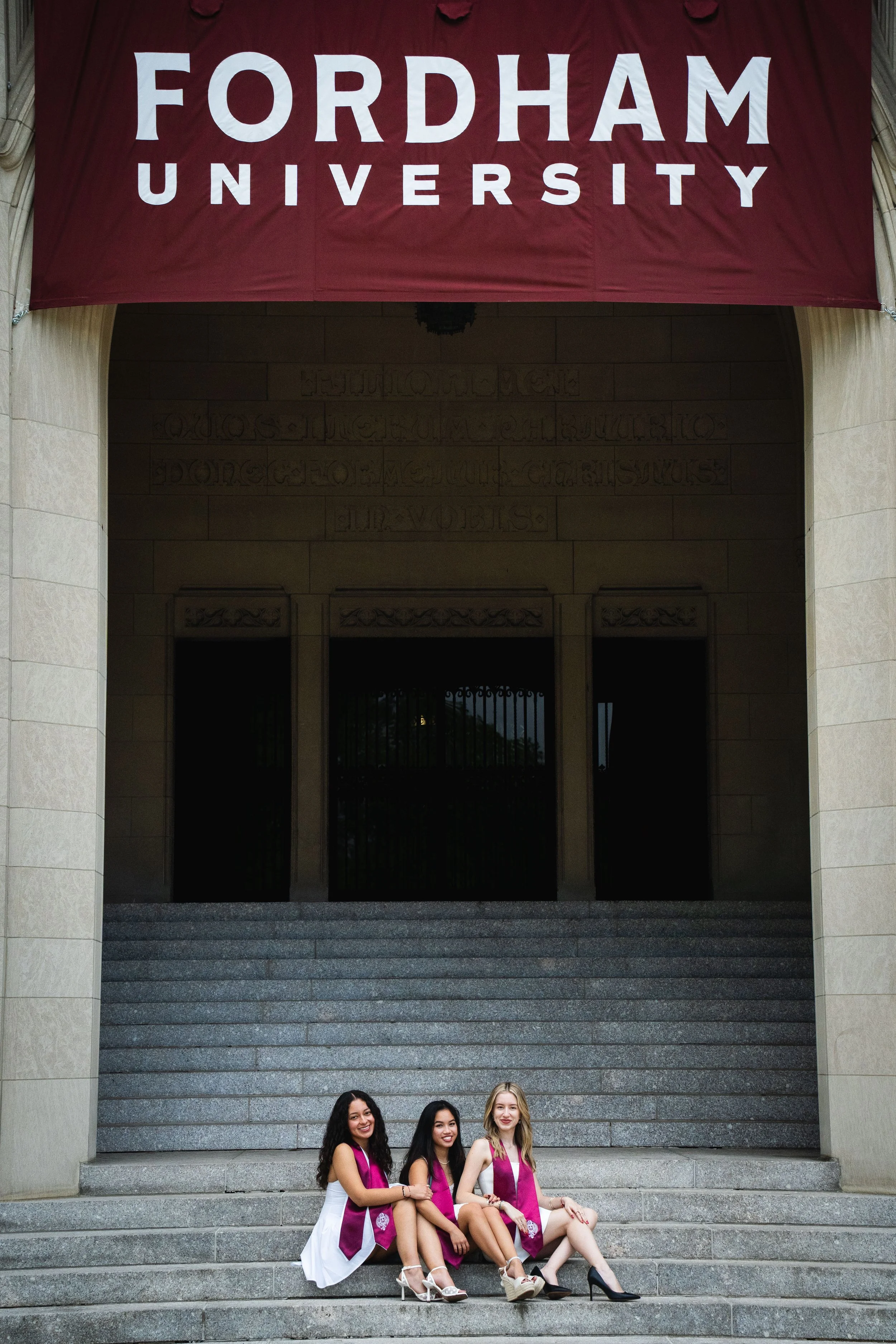 Three women sitting on the steps in front of a building at Fordham University, wearing white dresses and maroon sashes, smiling for the photo.