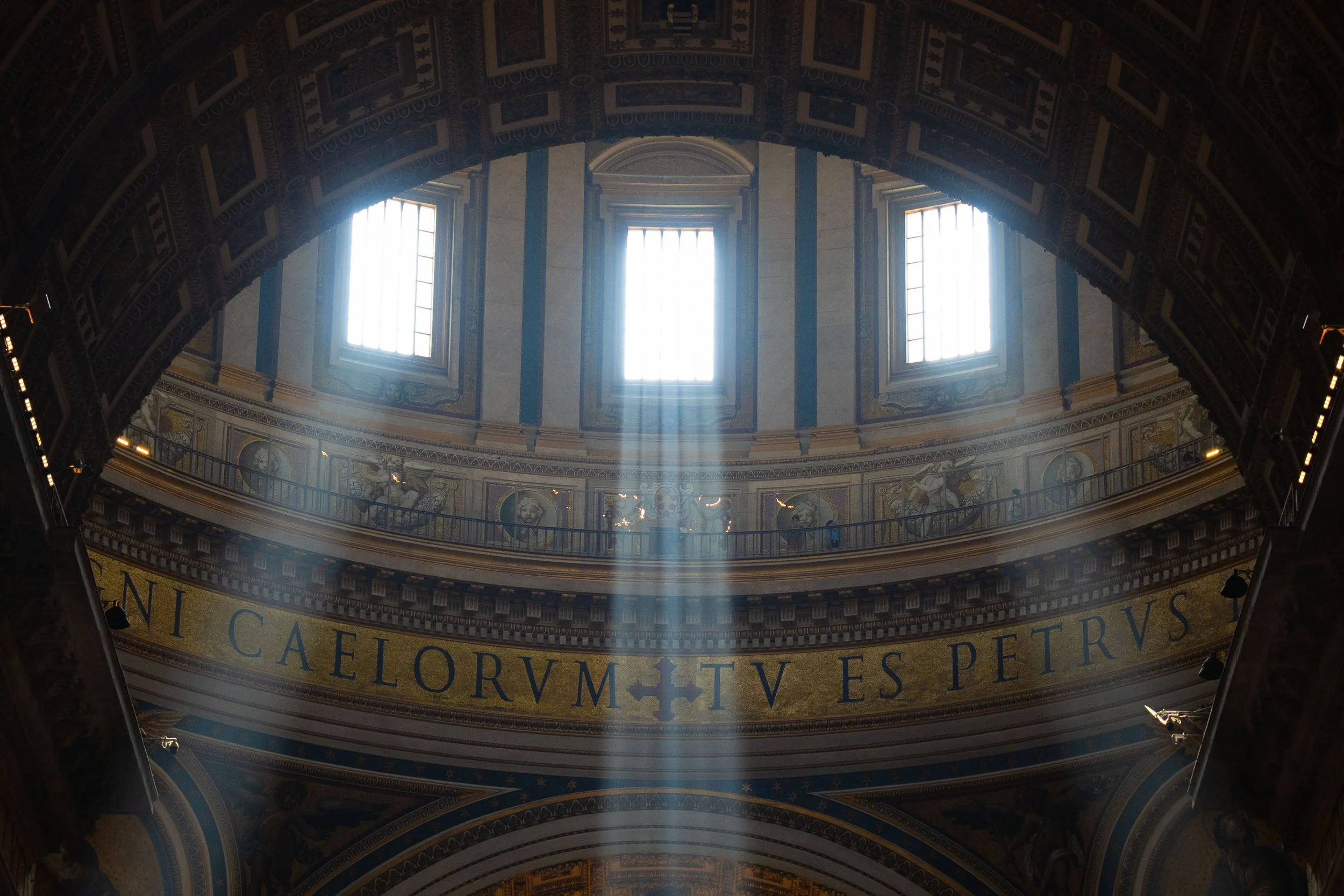 Inside view of a domed ceiling with three high windows allowing sunlight to stream through. The ceiling features intricate artwork and decorative elements, with Latin-looking text on a band around the interior.