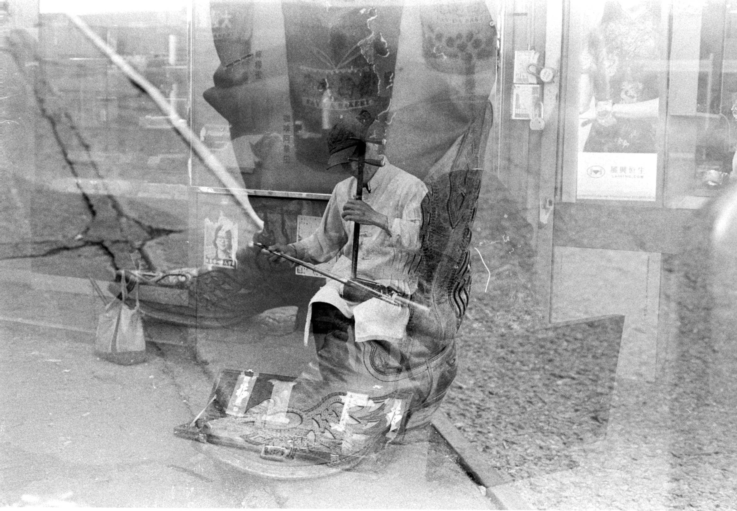 Reflective black-and-white photo of a person sitting outdoors, holding a fishing rod. The person is wearing a white jacket and dark pants with a striped pattern. Visible on the glass is the faint reflection of a roadside scene with cars and a street sign, and the background suggests a wooded area.