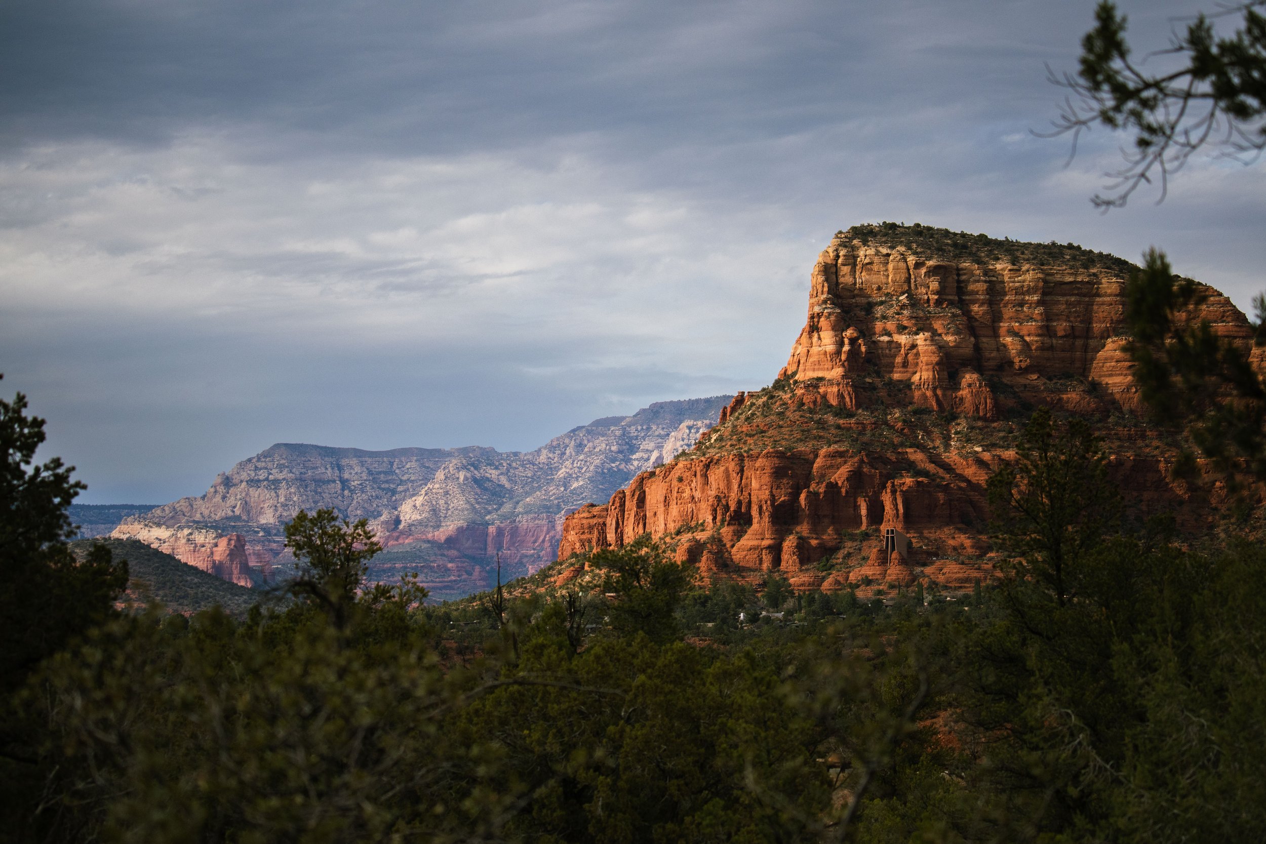 Scenic view of large red rock formations in a canyon under a cloudy sky, with trees and bushes in the foreground.