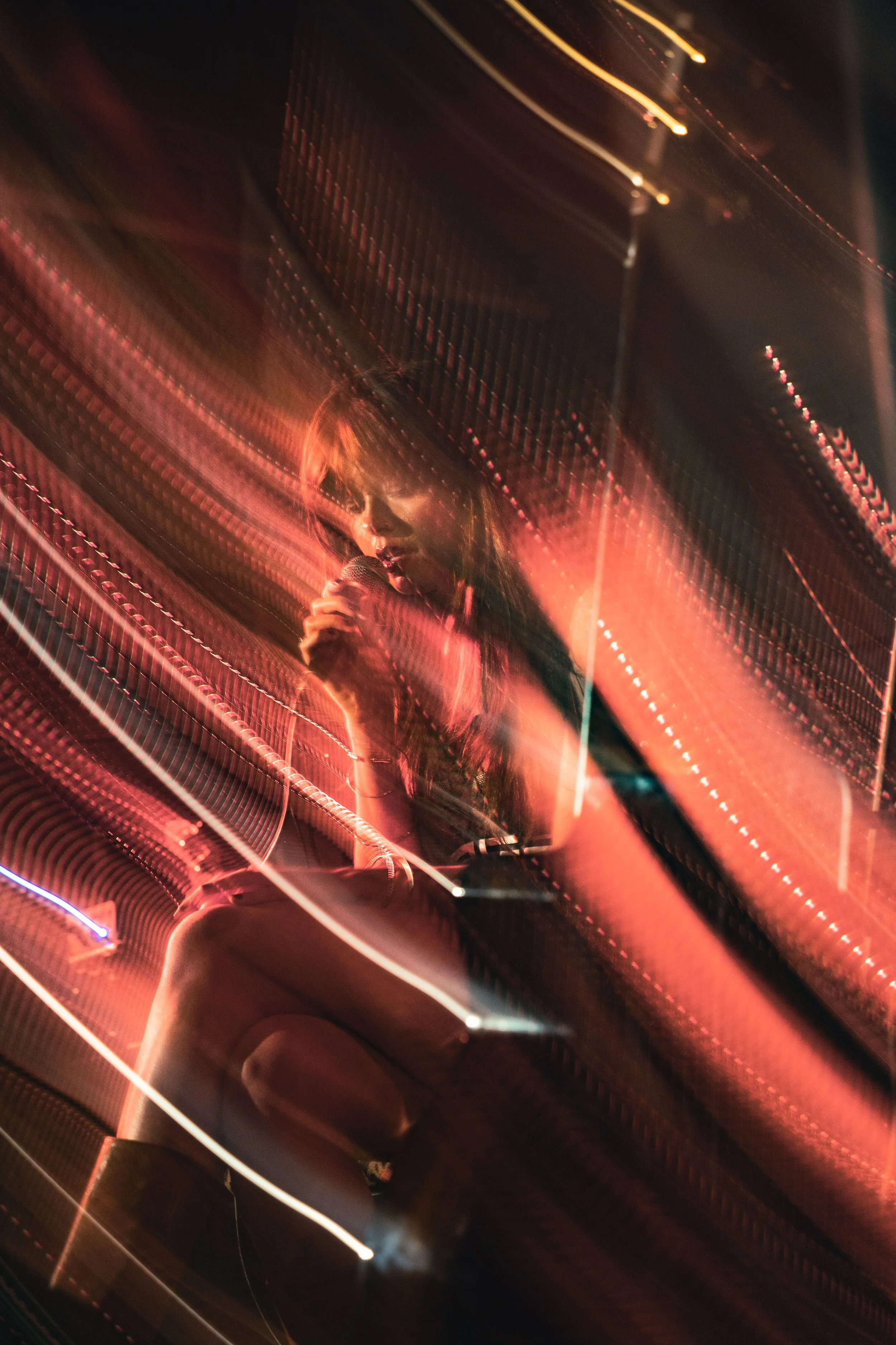 A woman singing into a microphone seen through a glass window with a reflective surface, creating an abstract, streaked effect with red and orange lighting.