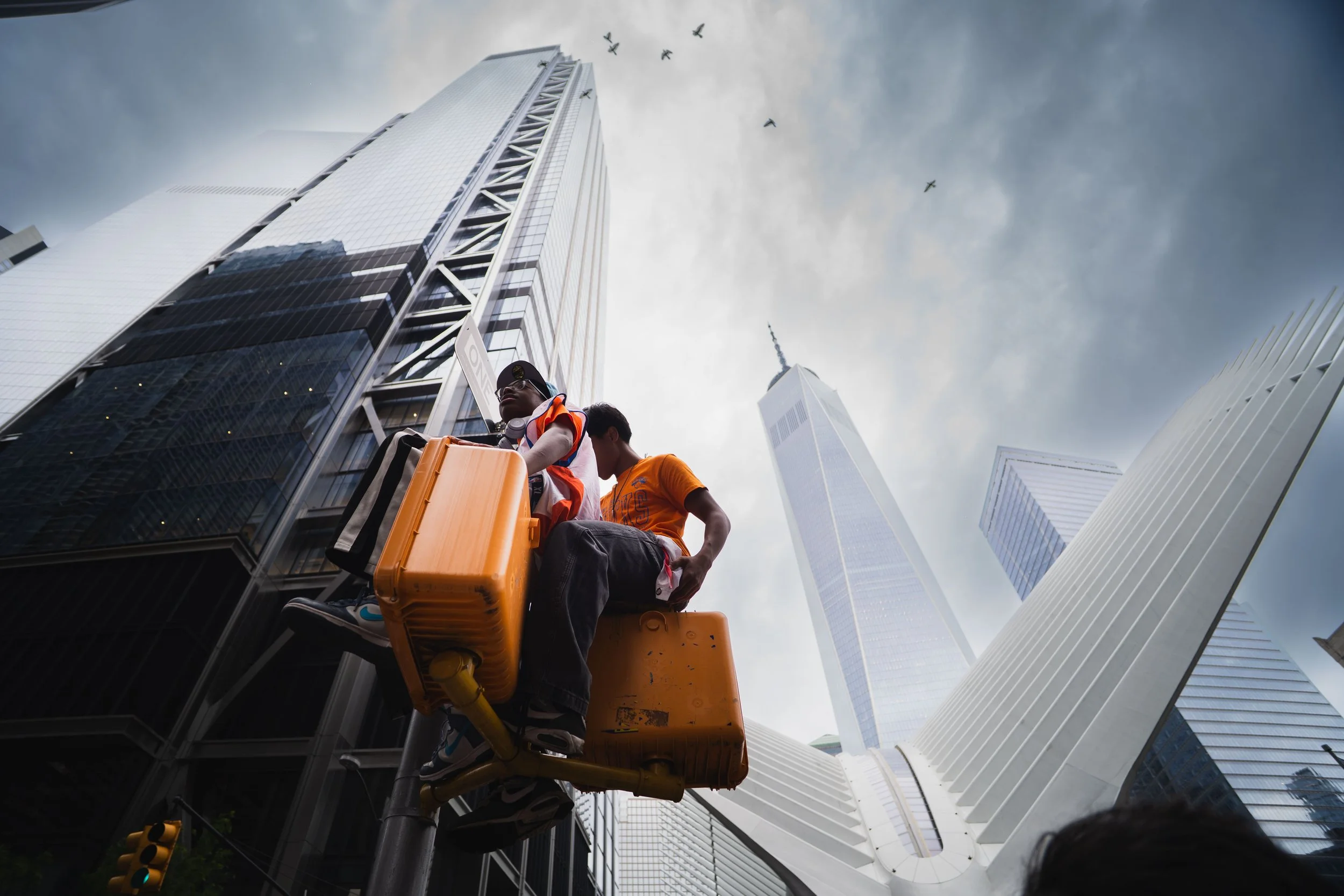 Two city workers on a yellow elevated work platform inspecting tall modern skyscrapers with glass facades in an urban cityscape under cloudy skies, with birds flying overhead.