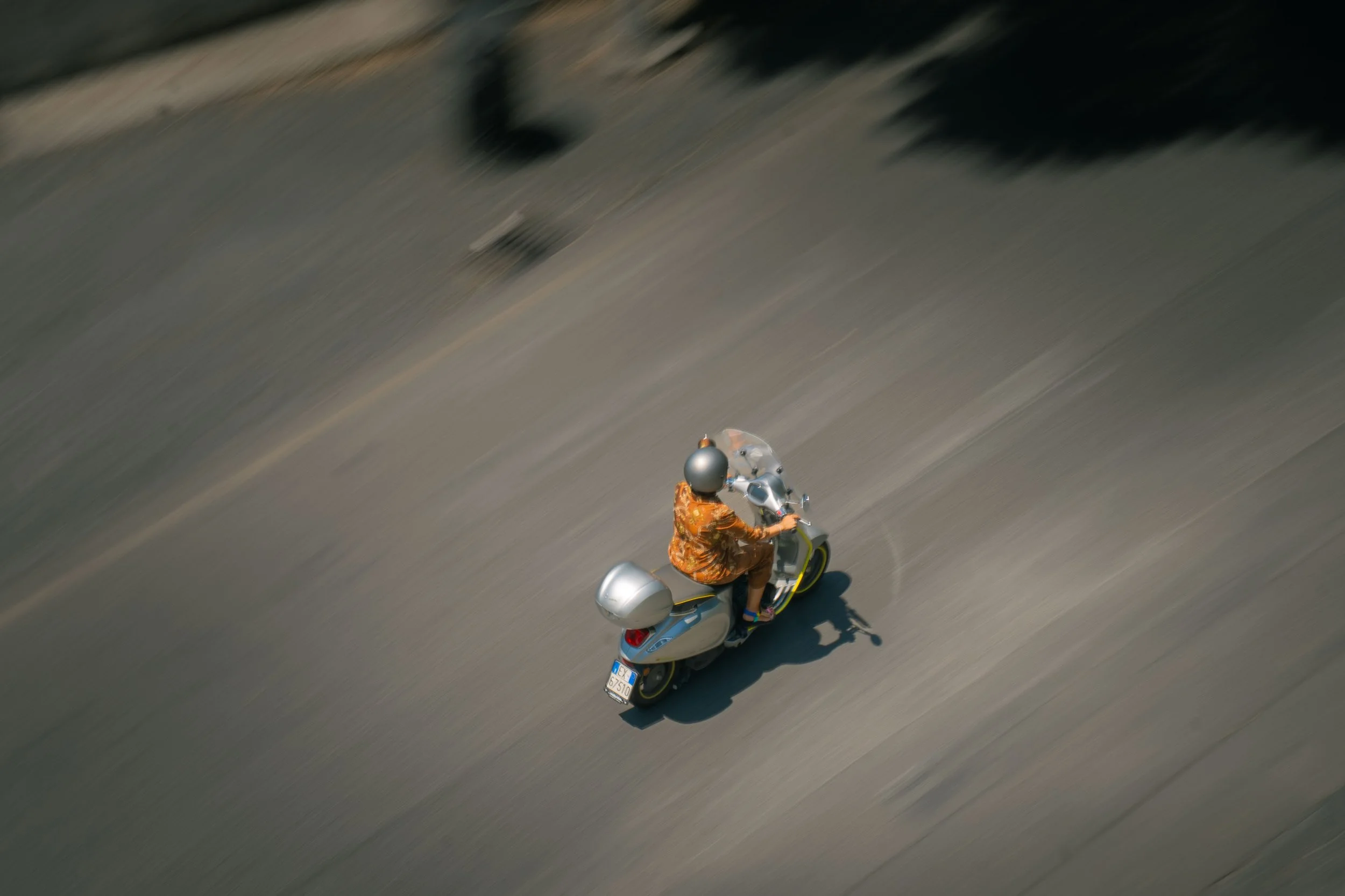 A person riding a silver scooter on a paved road, wearing a helmet and orange patterned clothing, with a blurred background indicating motion.