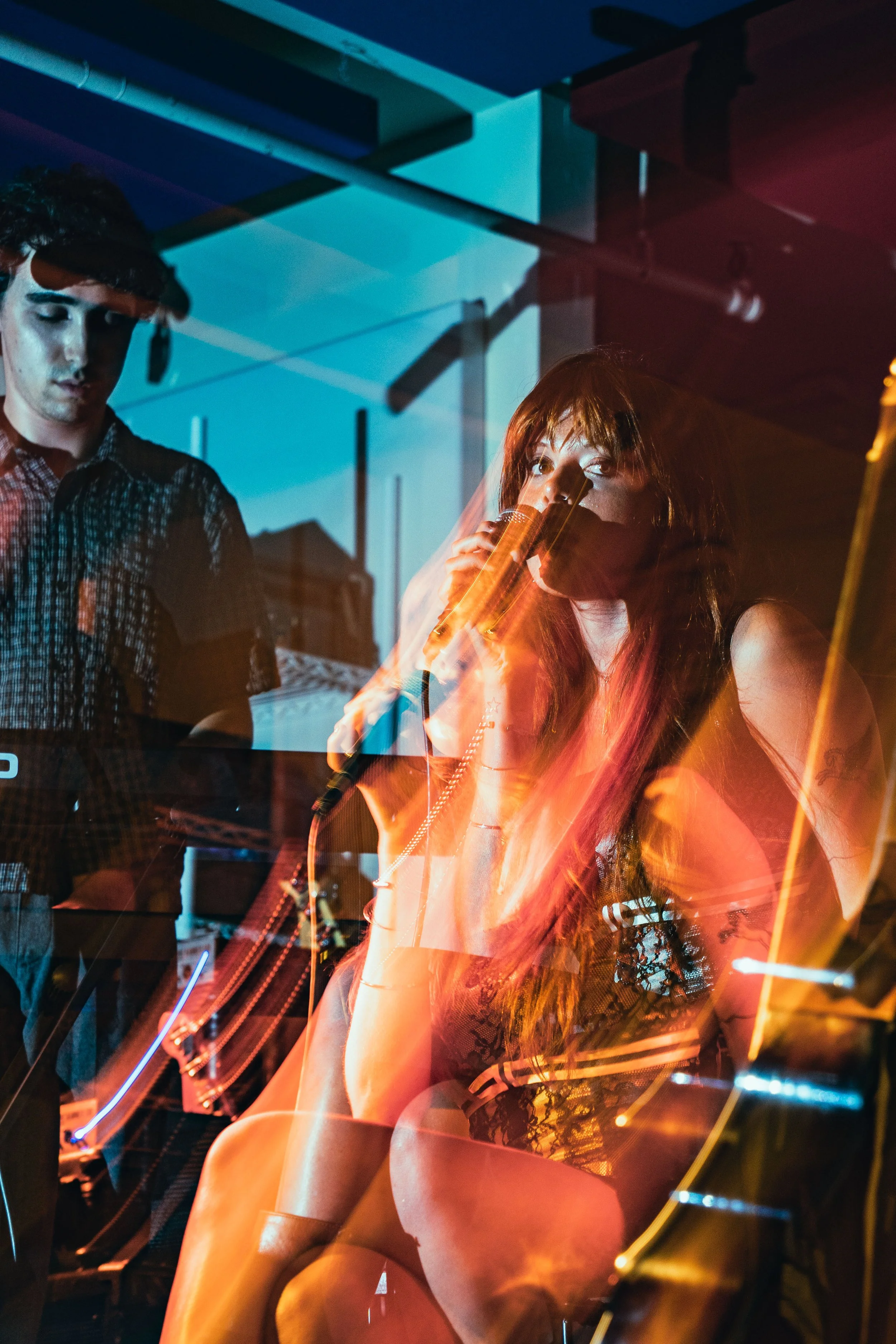 A woman with long red hair is sitting and singing into a microphone, with one leg crossed over the other. She is wearing a black lace top. Behind or reflection in glass showing a man with dark curly hair in a checkered shirt looking down, during a ni