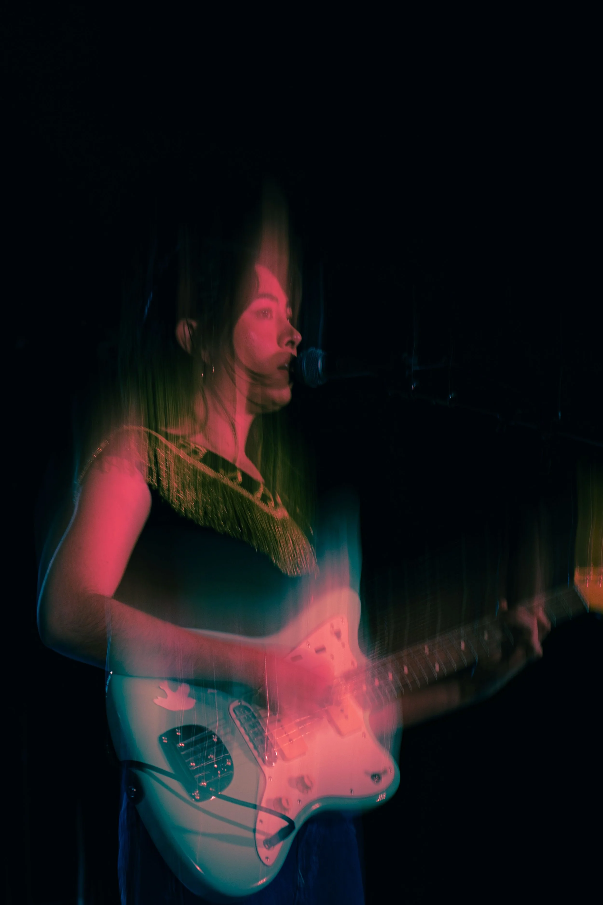 A woman singing into a microphone while playing an electric guitar on a dark stage.