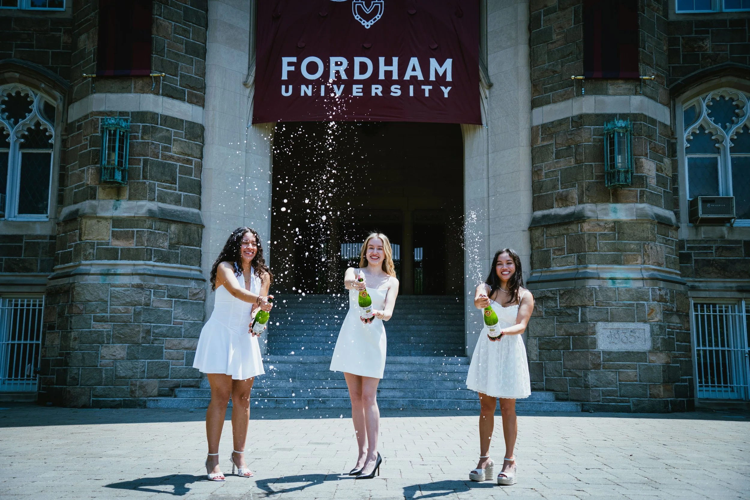 Three women in white dresses celebrating graduation outside Fordham University, spraying champagne.
