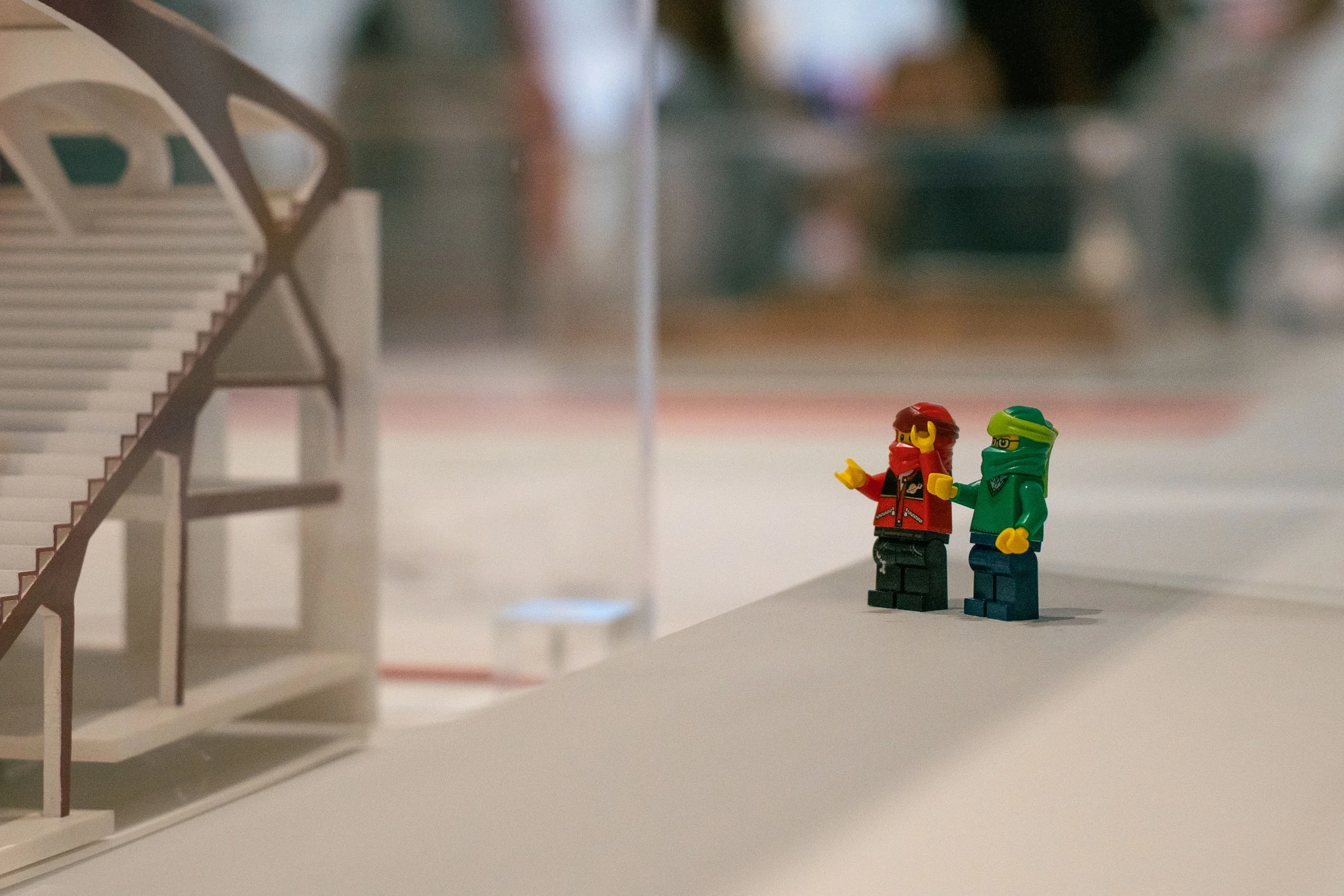 Close-up of two LEGO minifigures standing on a white surface, one with a red outfit and yellow face mask, the other with a green outfit and black glasses, with a blurred staircase structure in the background.