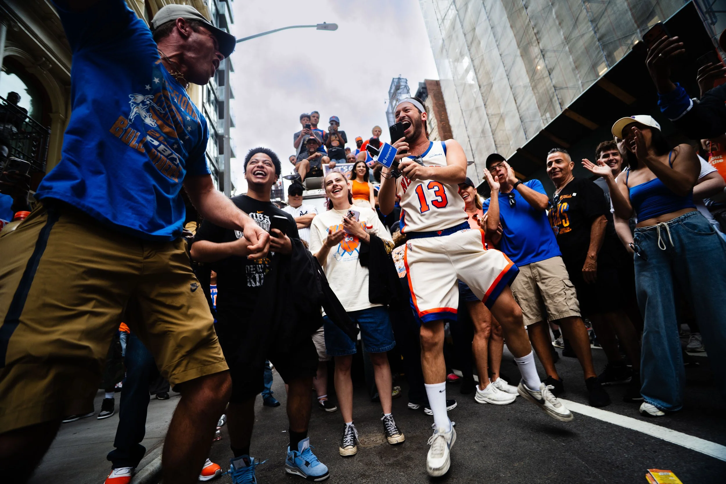 A lively street scene with a group of diverse people celebrating, some wearing New York Knicks jerseys, dancing and smiling amidst urban buildings and spectators.