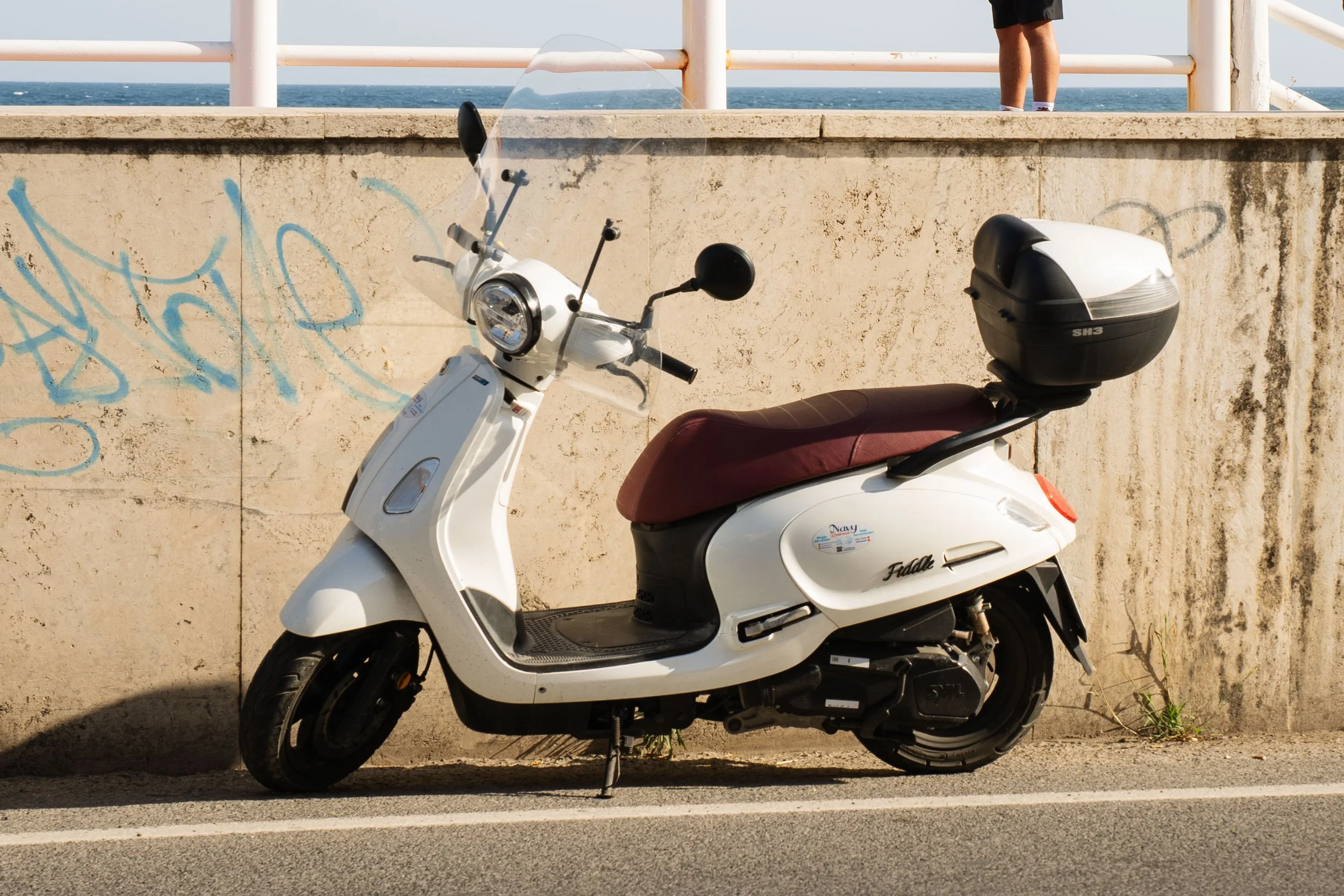 White scooter with a brown seat parked against a graffiti-covered beige wall near the beach. The ocean is visible in the background with white railings and a person standing above the scooter.