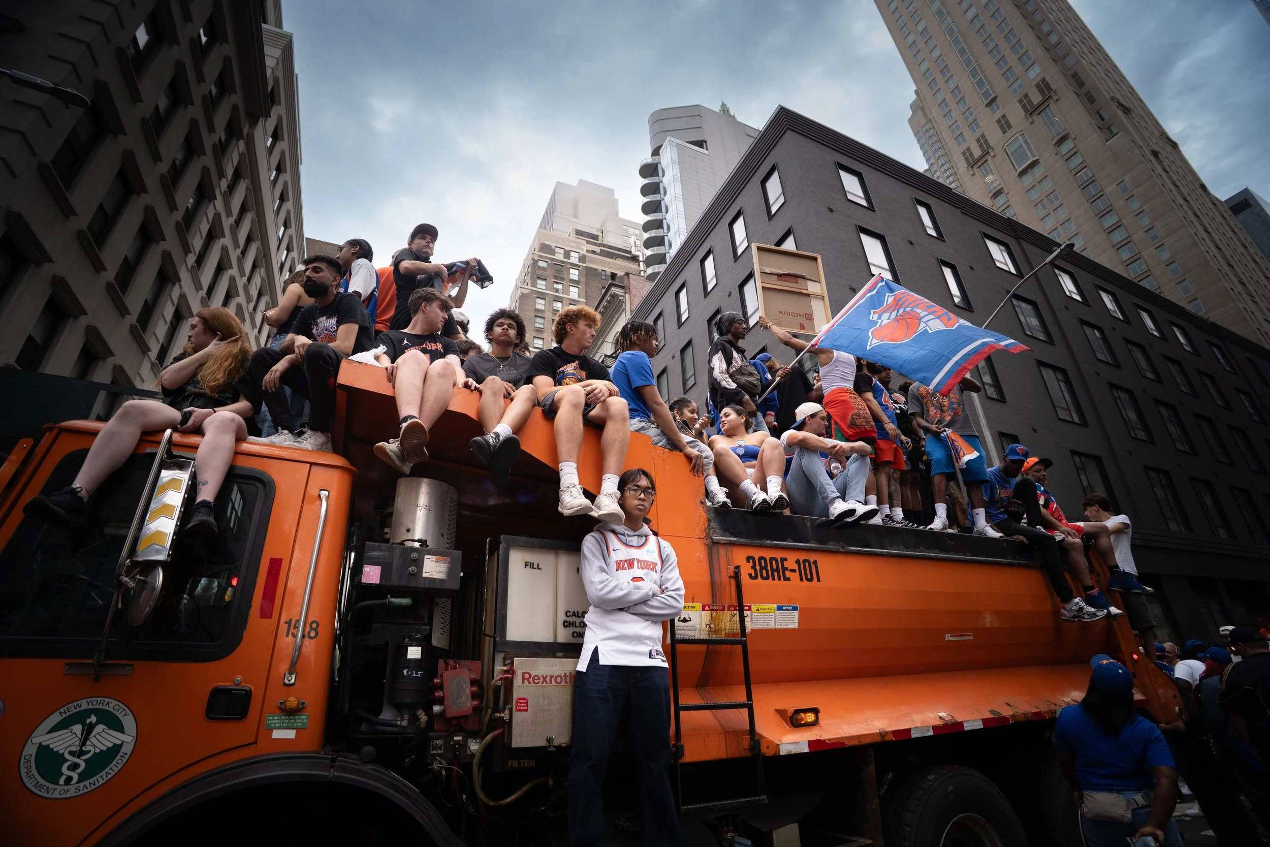 A group of people wearing New York Knicks apparel sitting and standing on top of an orange utility truck during a parade in an urban city street. Buildings and skyscrapers are visible in the background, with a partly cloudy sky overhead.