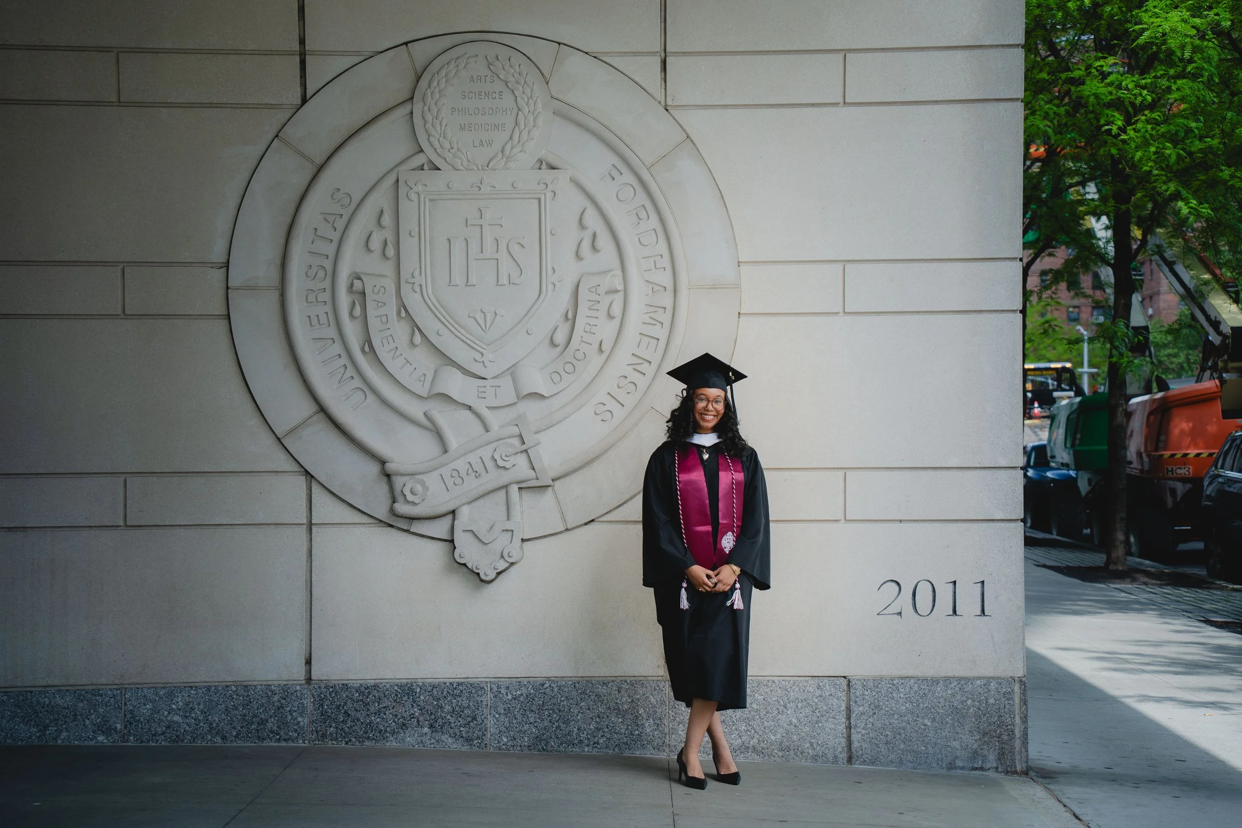 Graduate in cap and gown posing outdoors in front of university emblem on wall, with trees and city street in background.