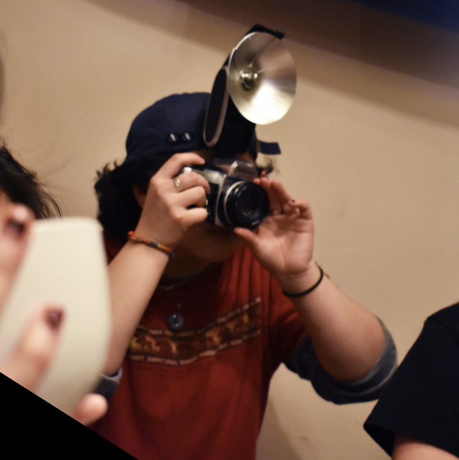 Person wearing a black cap, red shirt, and camera around their neck, taking a photo indoors with a camera.