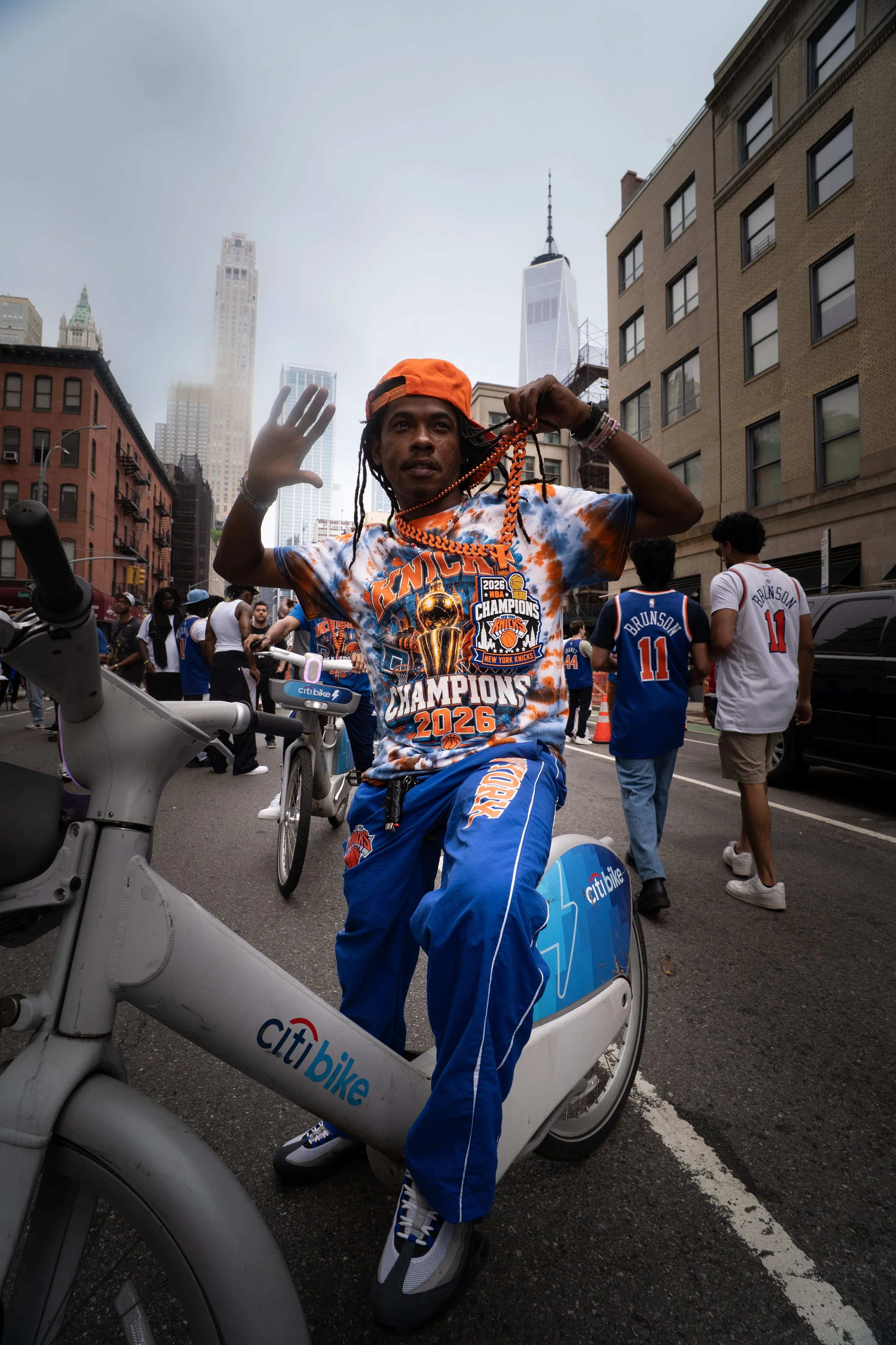 A young man with dreadlocks wearing a bright orange cap, tie-dye Knicks championship t-shirt, and blue athletic pants is sitting on a Citi Bike, holding colorful necklaces and waving with his left hand in a busy city street with skyscrapers and peopl