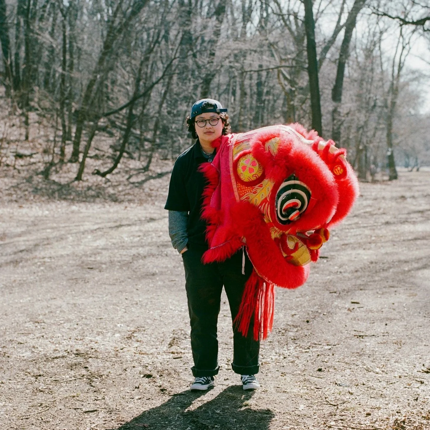A person with glasses, a black cap, and casual clothing standing outdoors on a dirt path, holding a red and gold Chinese lion dance costume head, with leafless trees in the background.