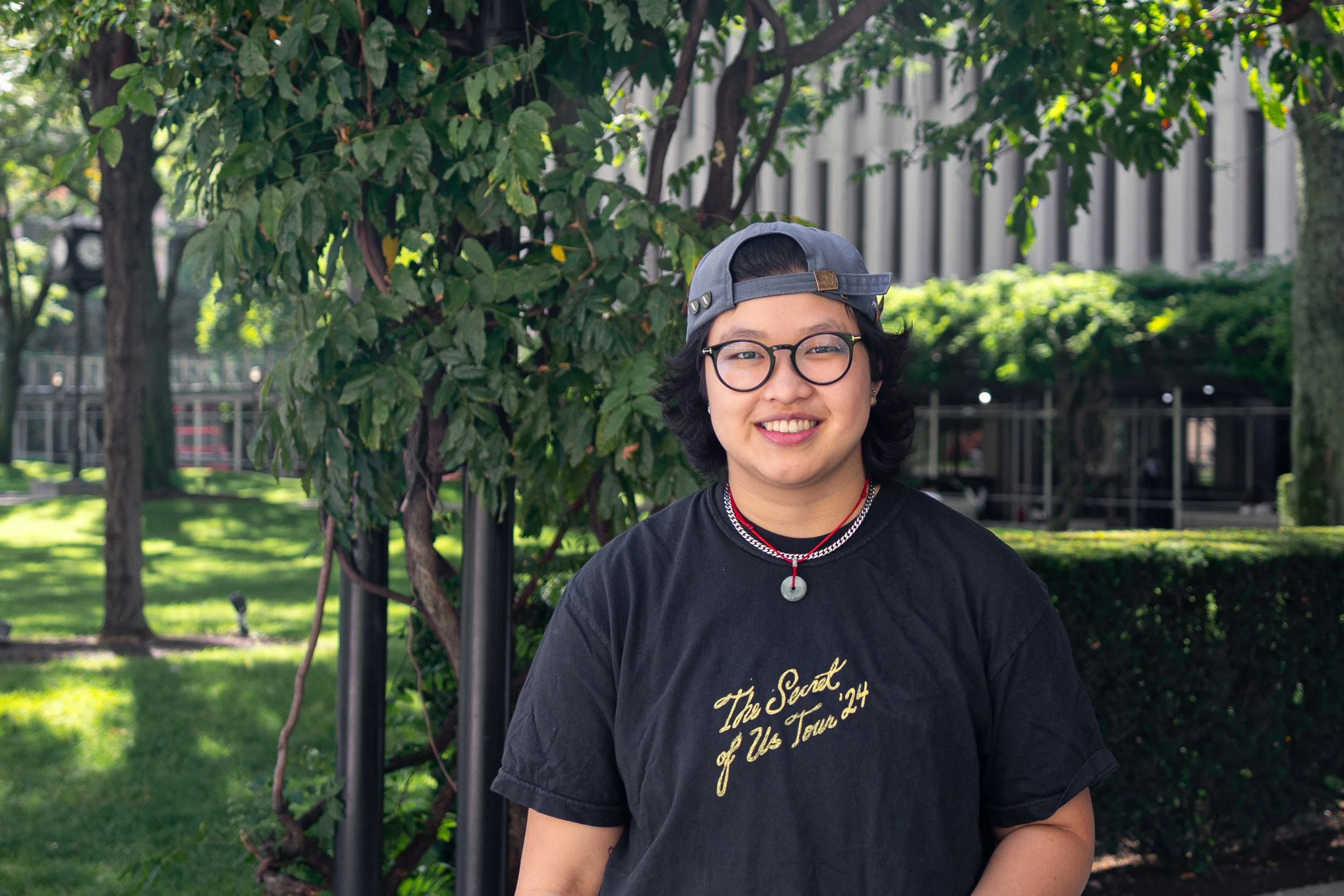 A person with short black hair, wearing glasses, a gray backward cap, and a black T-shirt with yellow writing, standing outdoors in front of green trees and grass, smiling at the camera.