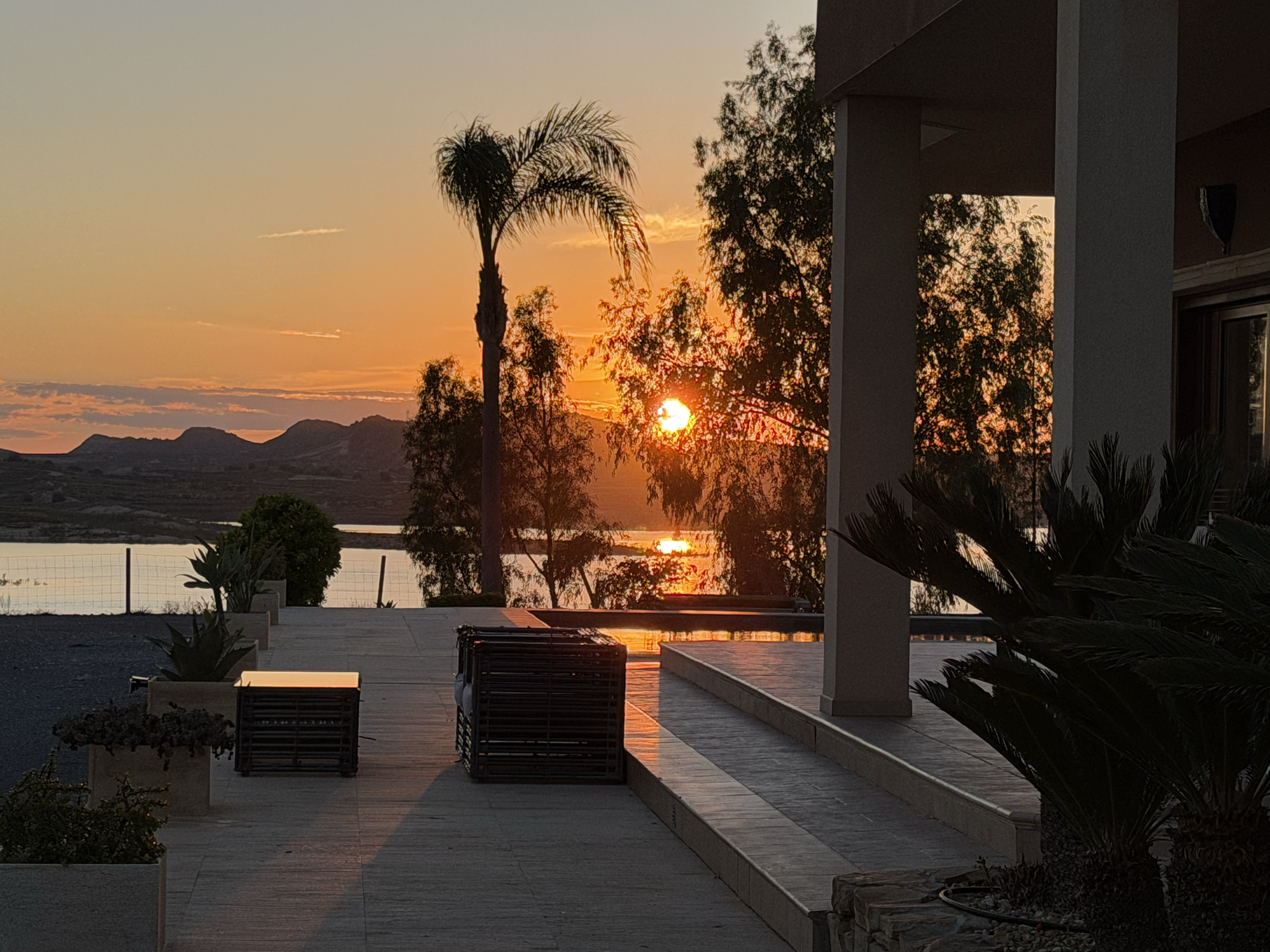 Sunset over a patio with outdoor seating, potted plants, palm trees, and mountains in the background.