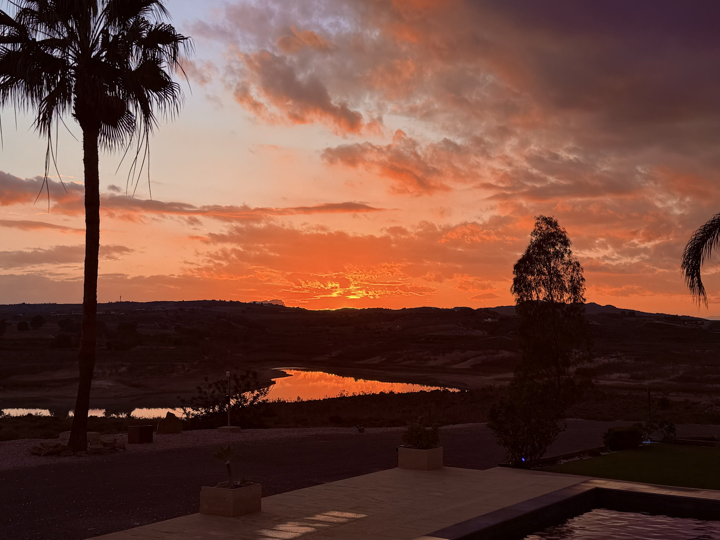 A sunset with orange and pink clouds over a landscape with palm trees and a small body of water.