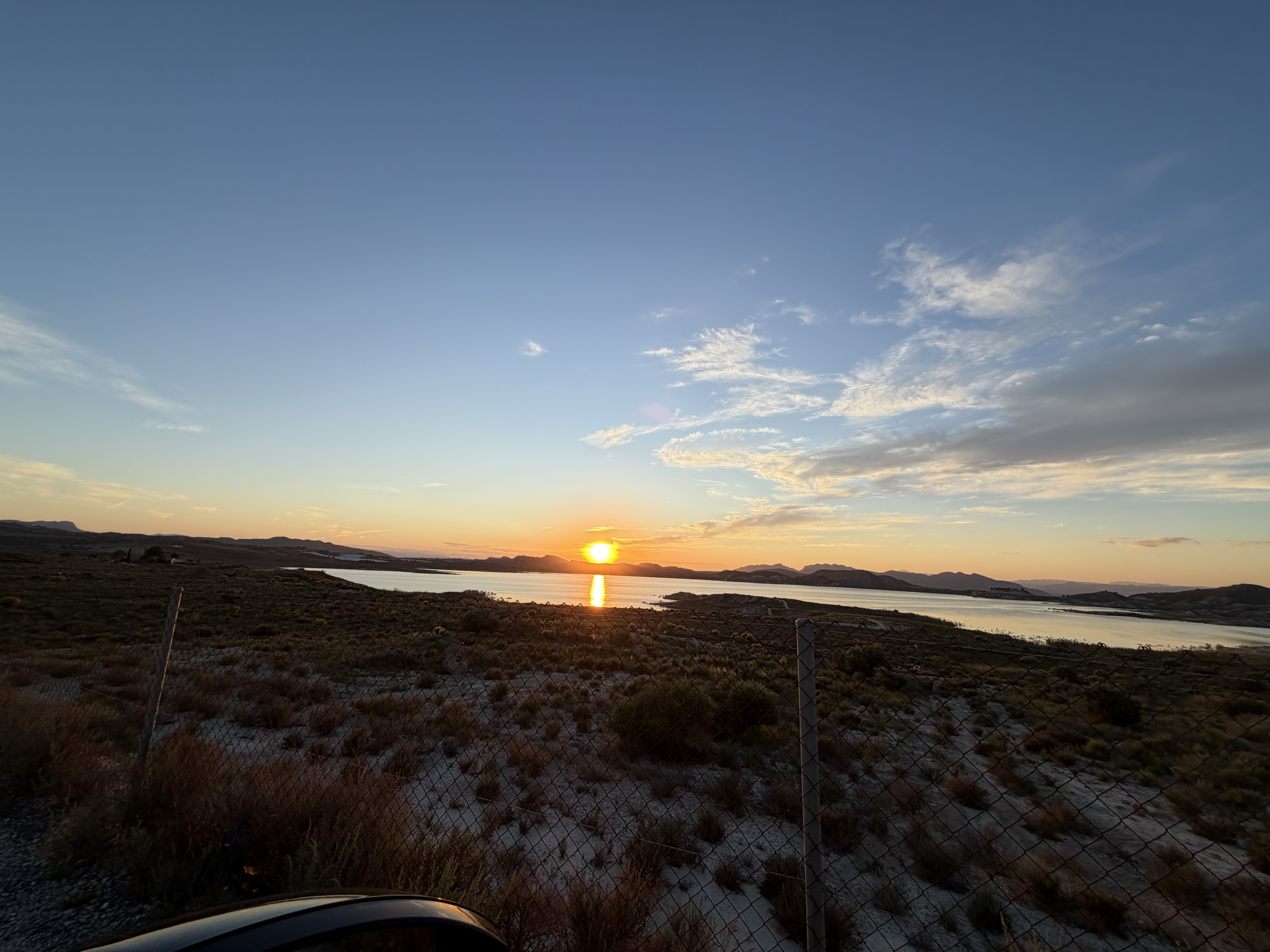 A landscape view of a sunset over a body of water with distant hills and a sky partly cloudy.