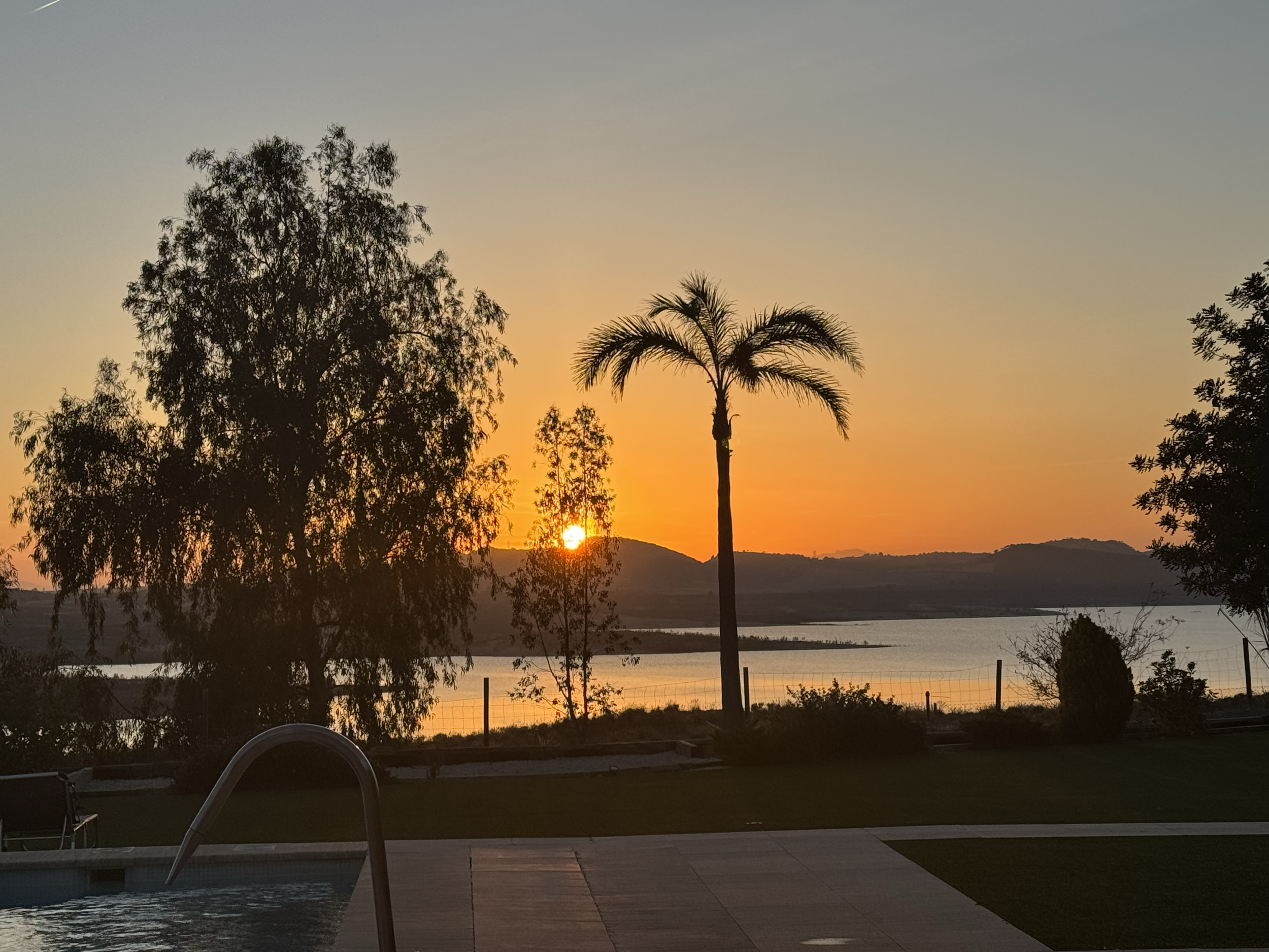 Sunset view over a lake with trees in the foreground, a fence, and a swimming pool, during twilight.