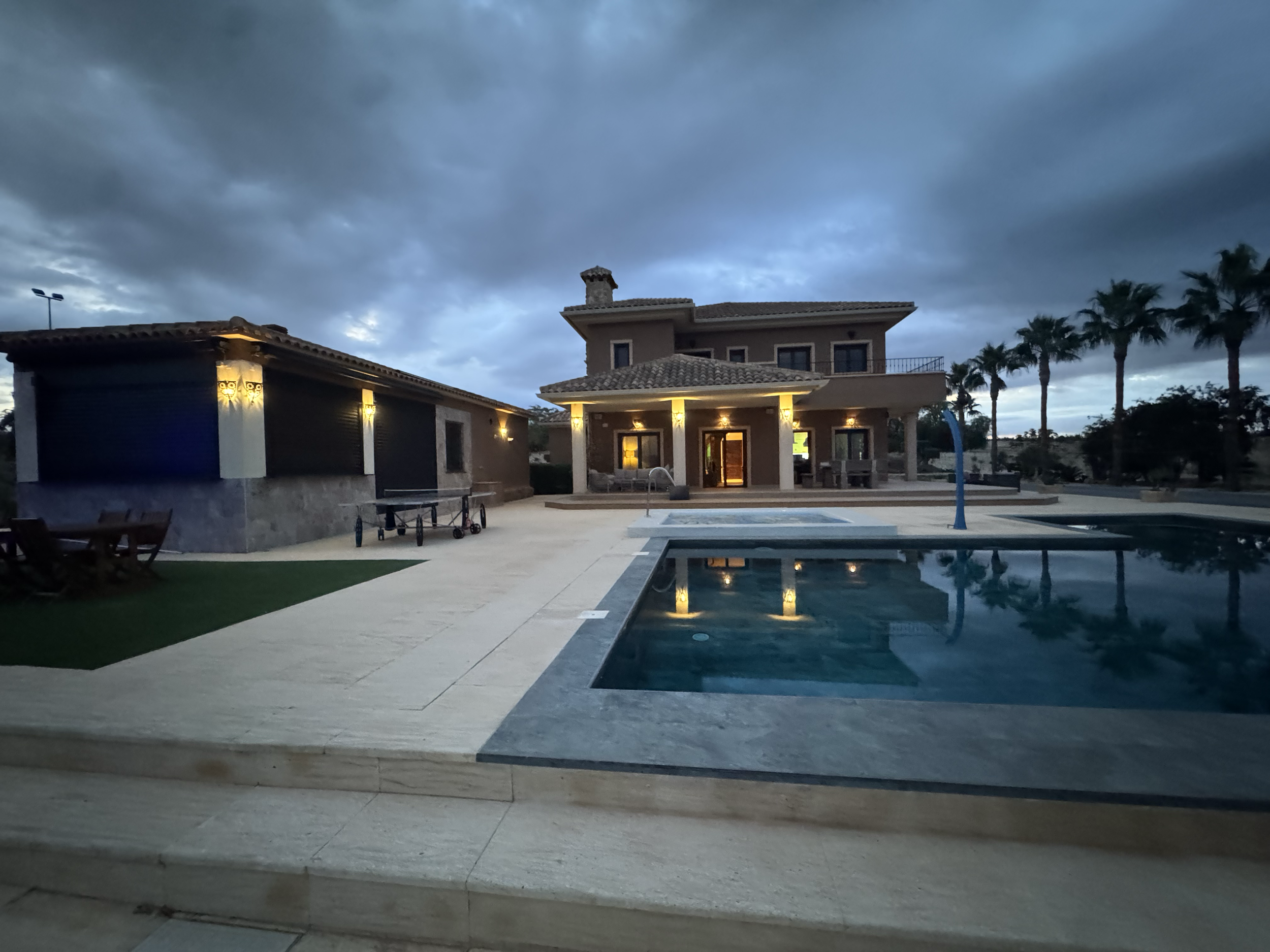 A backyard with a swimming pool, a house with outdoor lighting, palm trees, and cloudy skies at dusk.