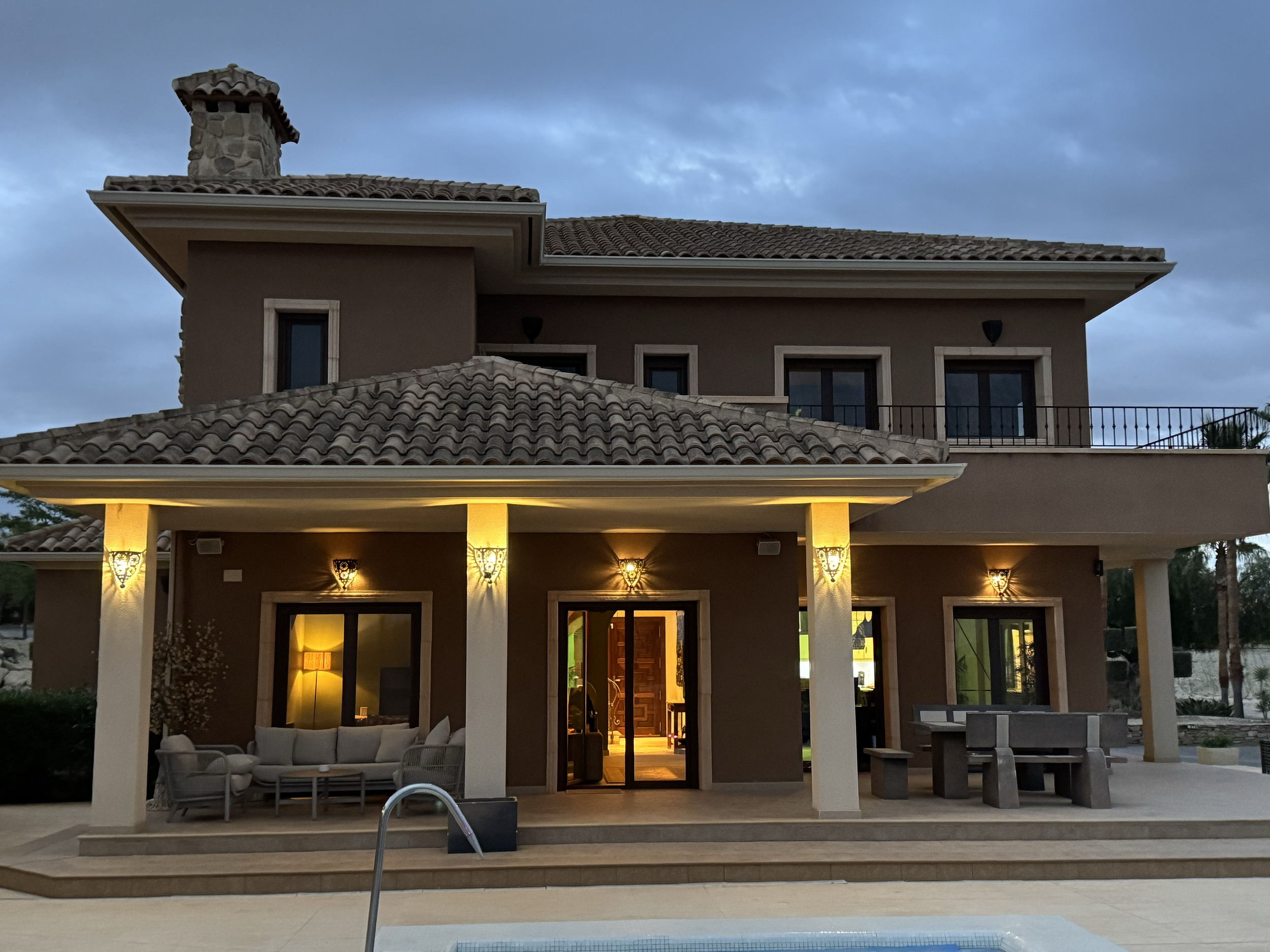 A two-story house with a tiled roof, exterior lighting, large glass doors, a patio with outdoor seating, and a swimming pool in the foreground during twilight.