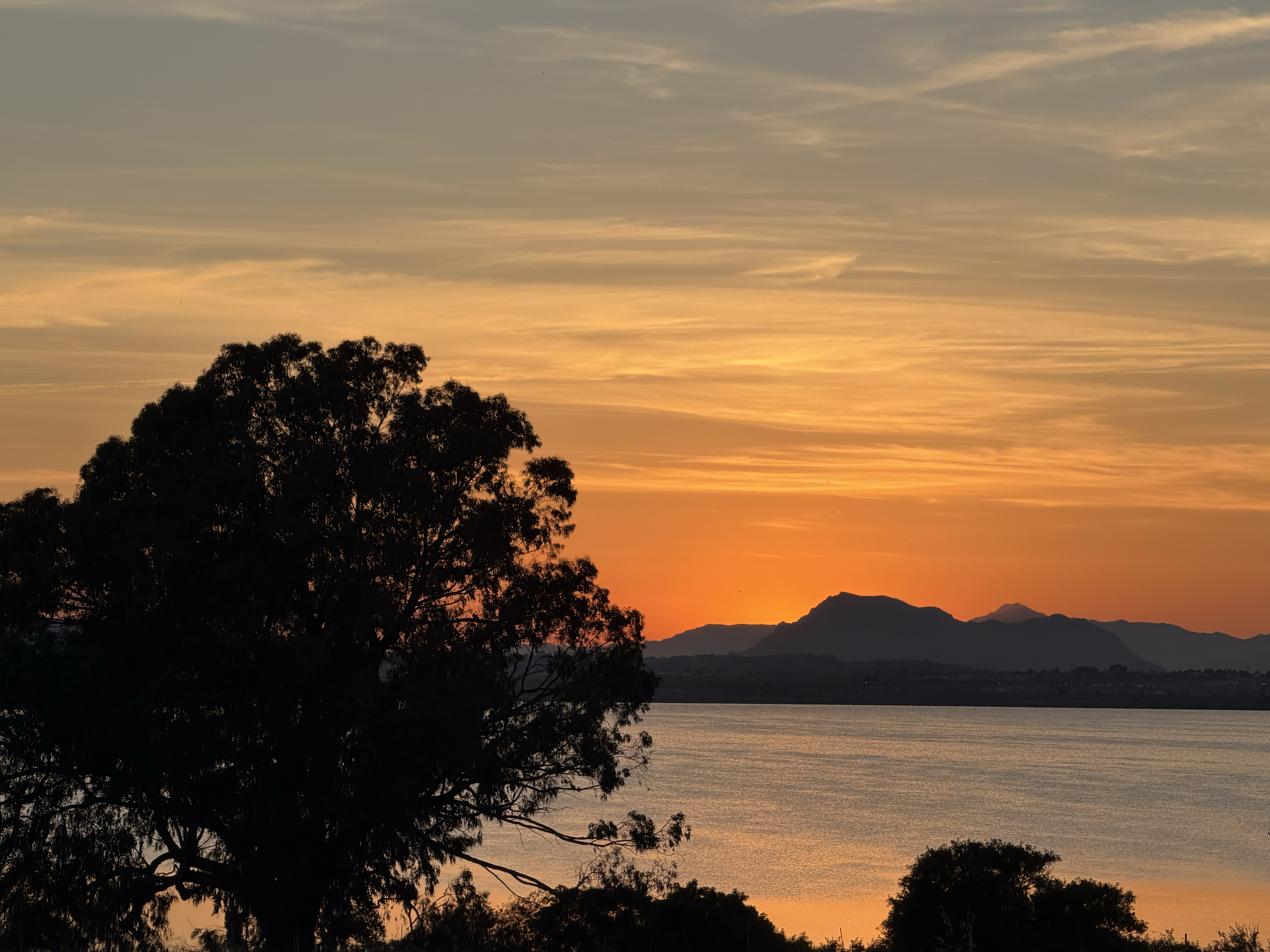 A sunset over a body of water with silhouettes of trees in the foreground and mountains in the background, with a partly cloudy sky.