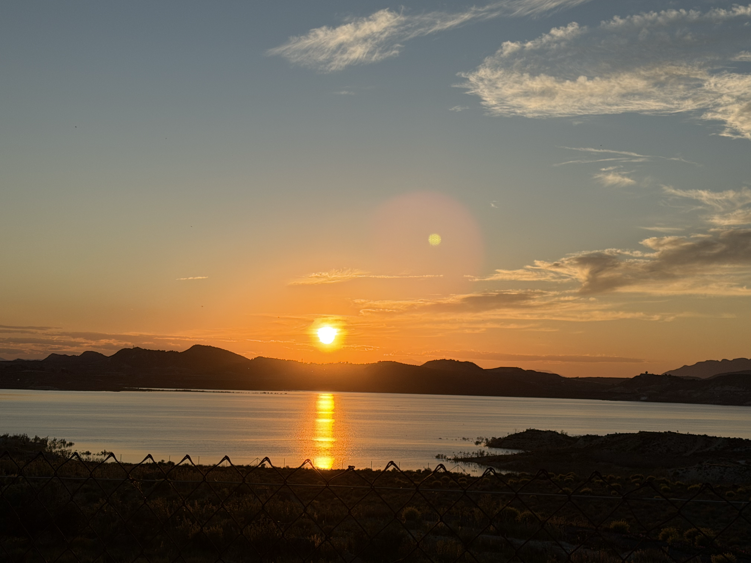 A sunset over a body of water with mountains in the background, clouds in the sky, and a chain-link fence in the foreground.