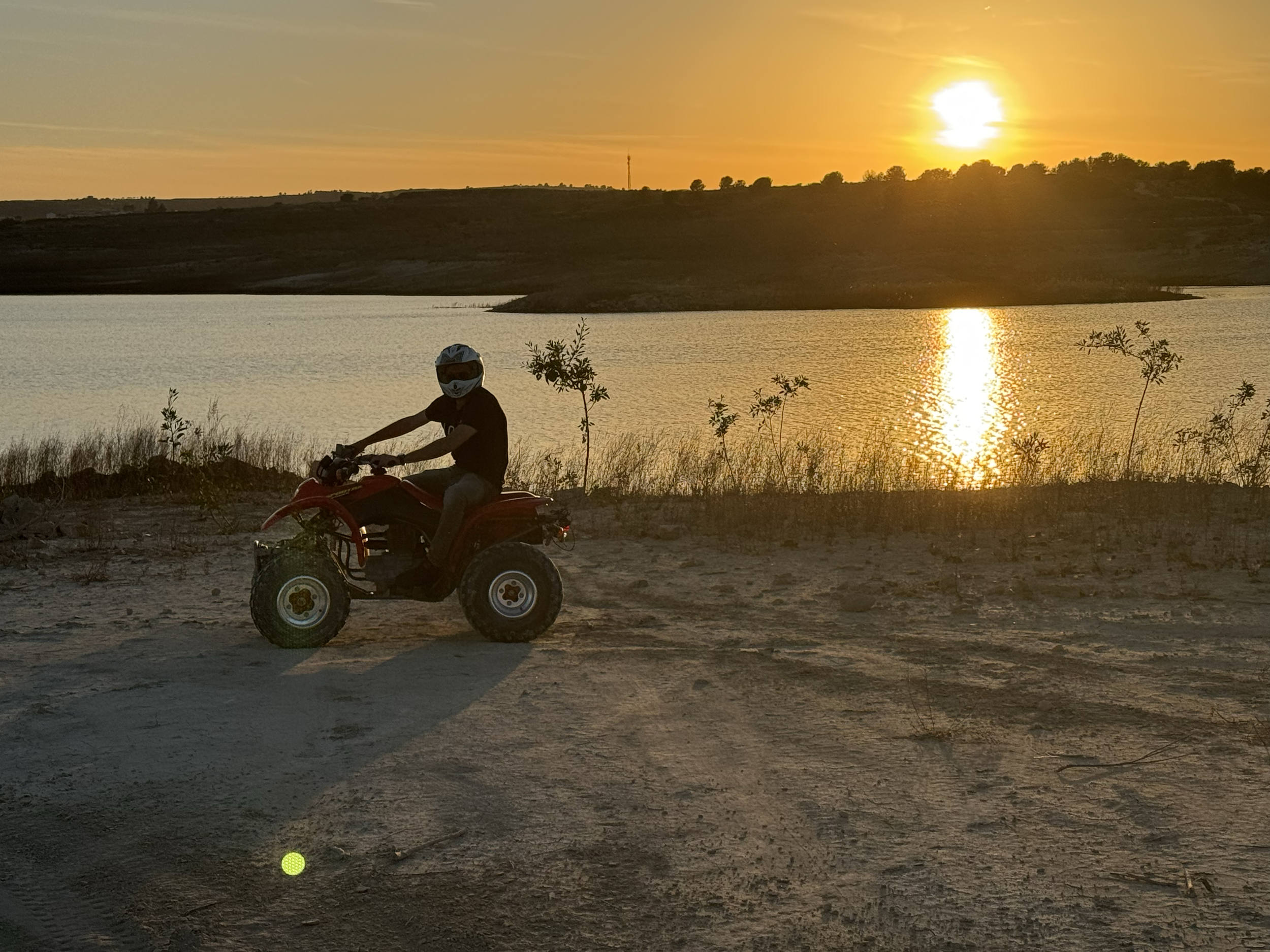 Person riding an all-terrain vehicle (ATV) near a body of water during a sunset, with trees and hills in the background.