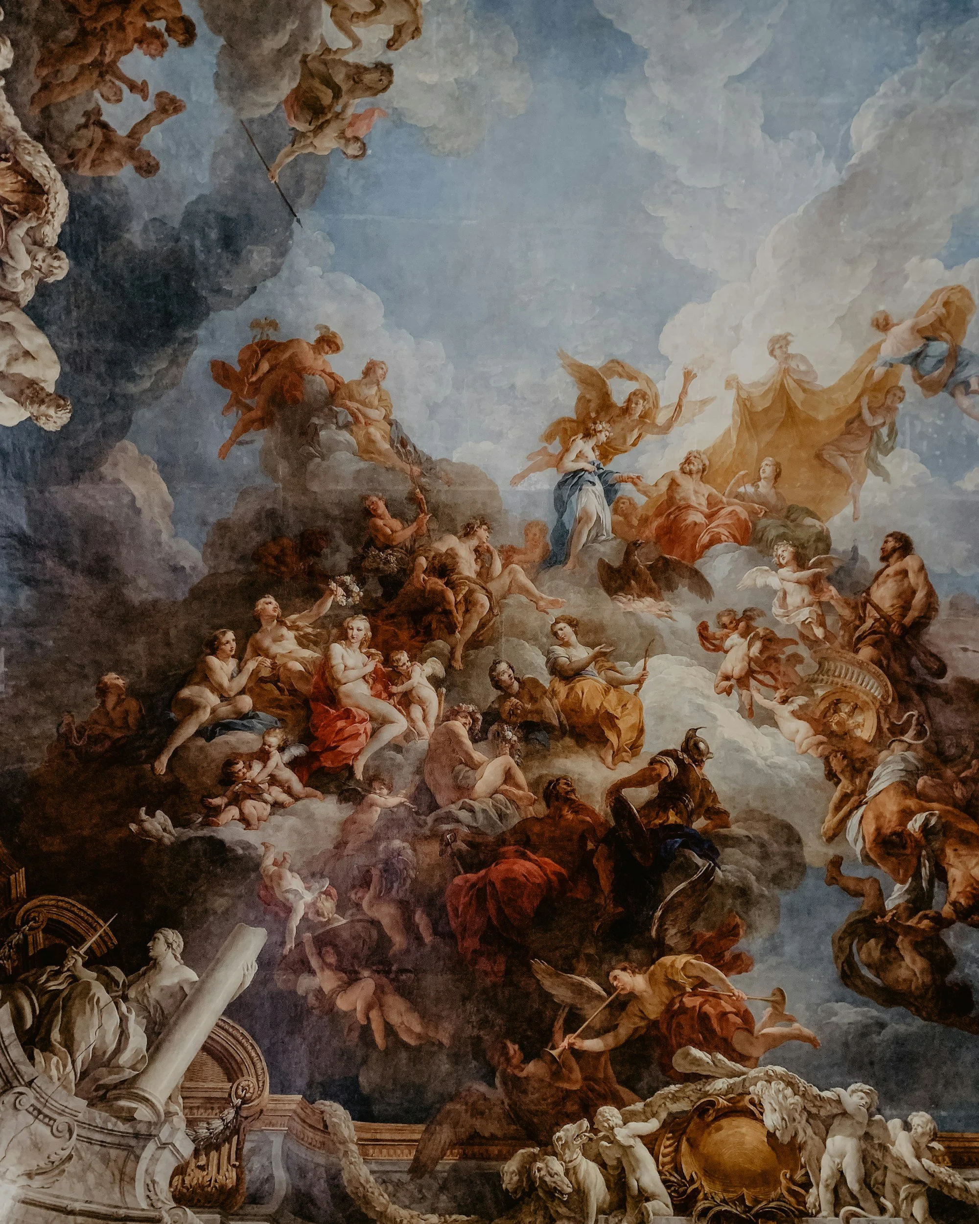 A detailed baroque ceiling painting depicting numerous mythological and cherubic figures surrounded by clouds, with some figures holding objects and engaged in dynamic poses, set against a partly cloudy sky background.