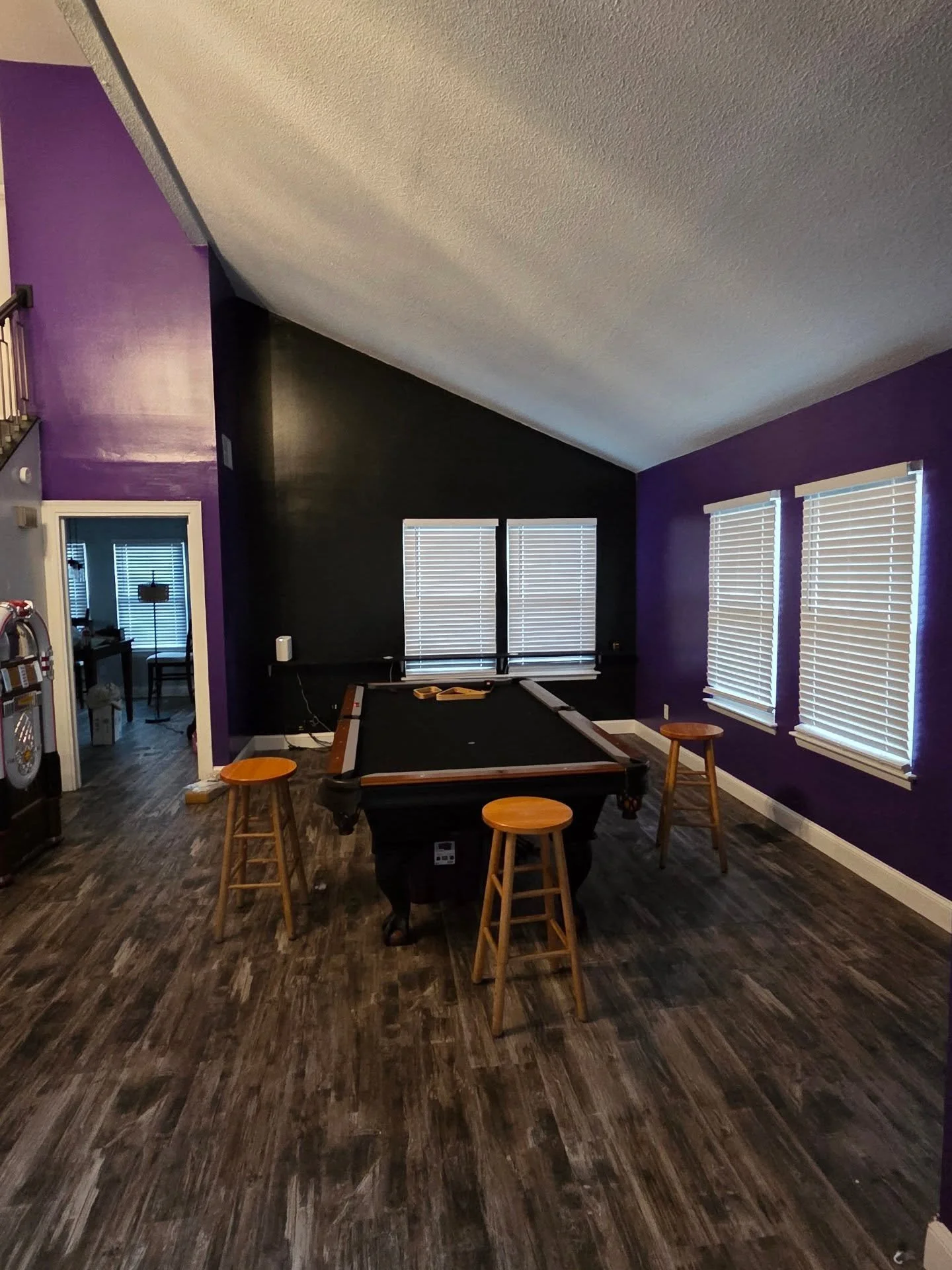 Living room with purple and black walls, three large windows with blinds, dark wood floor, pool table in the center, three wooden stools around it, an open door revealing a dining area with a window and chair, and various objects on the walls and surfaces.