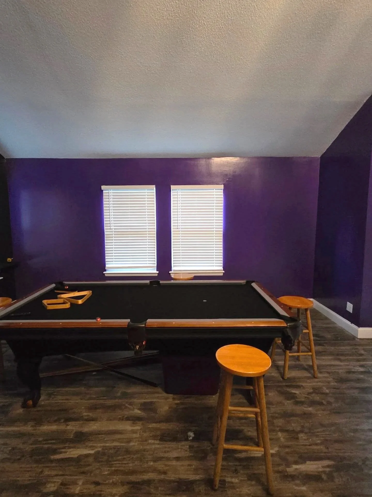 A room with purple walls, two windows with blinds, a pool table, small wooden stools, and a wooden floor.