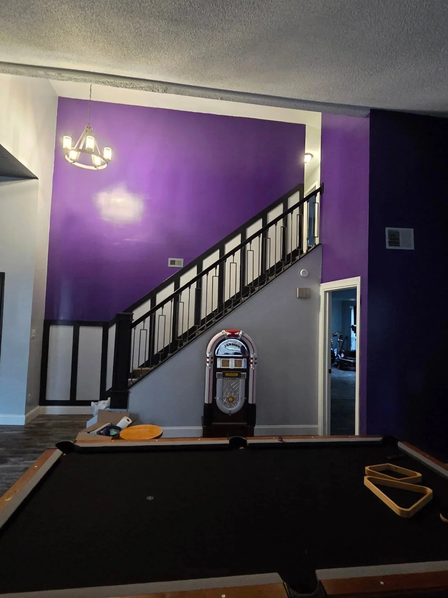 Interior view of a living room area with a purple accent wall, staircase with black railing, a hanging chandelier, a vintage jukebox, and a pool table in the foreground. Items and papers are scattered on the pool table.