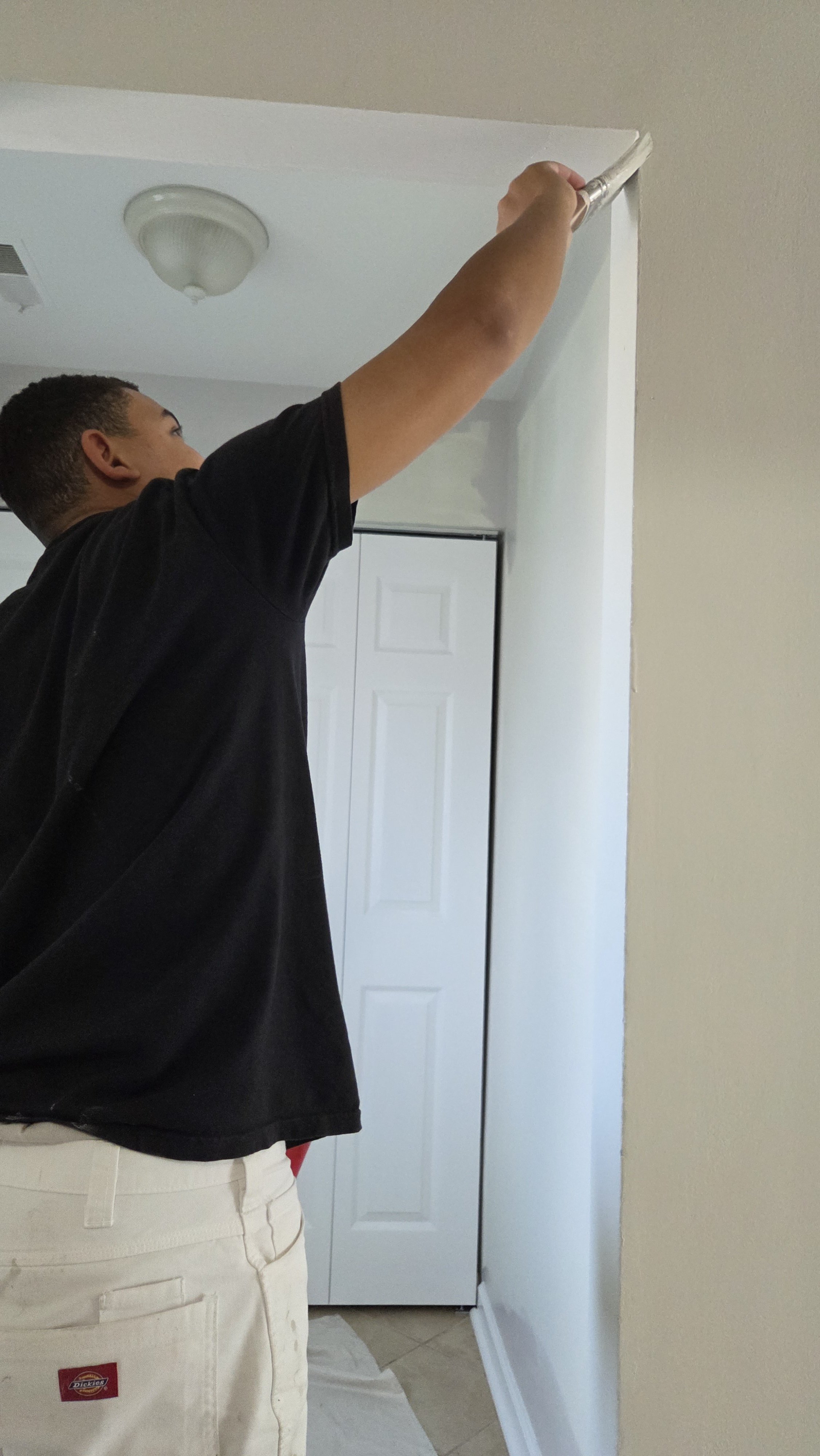 A person painting a ceiling with a paint roller in a room with a closet door, beige walls, and a ceiling light fixture.