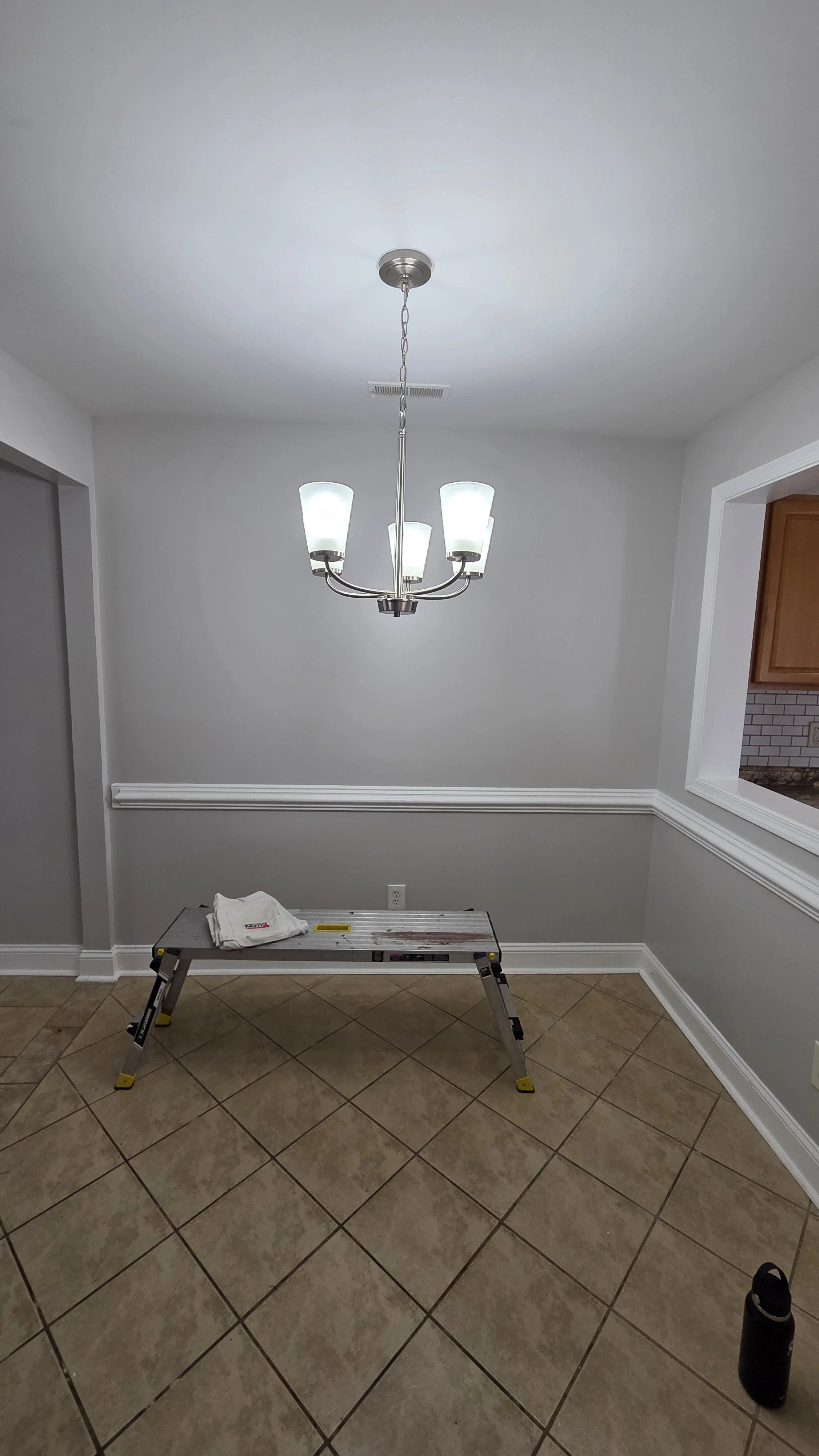 Empty dining room with a modern chandelier, tile floor, and a step ladder with a white towel on top. There is a partial view of a kitchen through a pass-through window to the right.