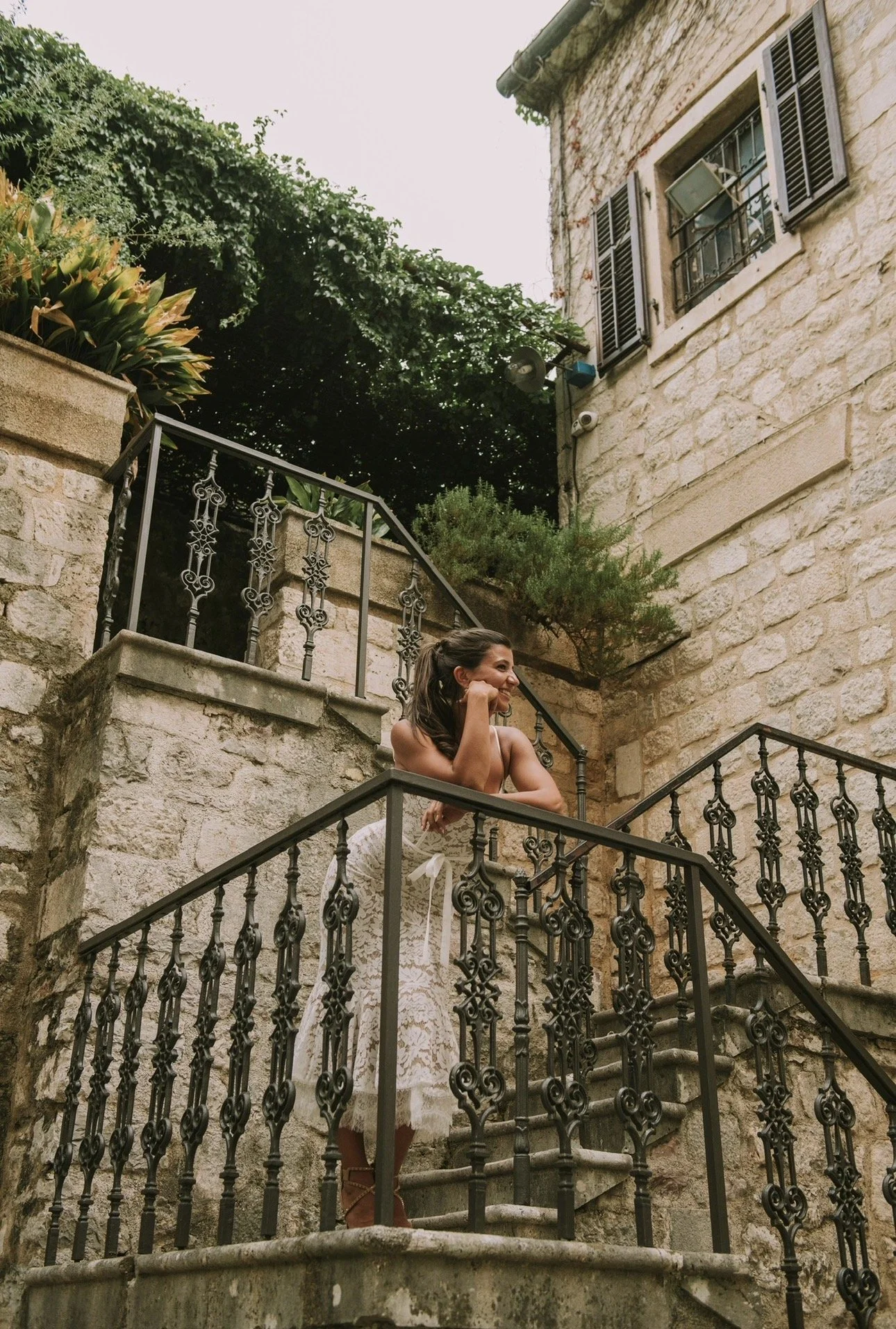 A woman standing on an outdoor stone staircase with ornate black metal railings, smiling, with buildings and greenery in the background.