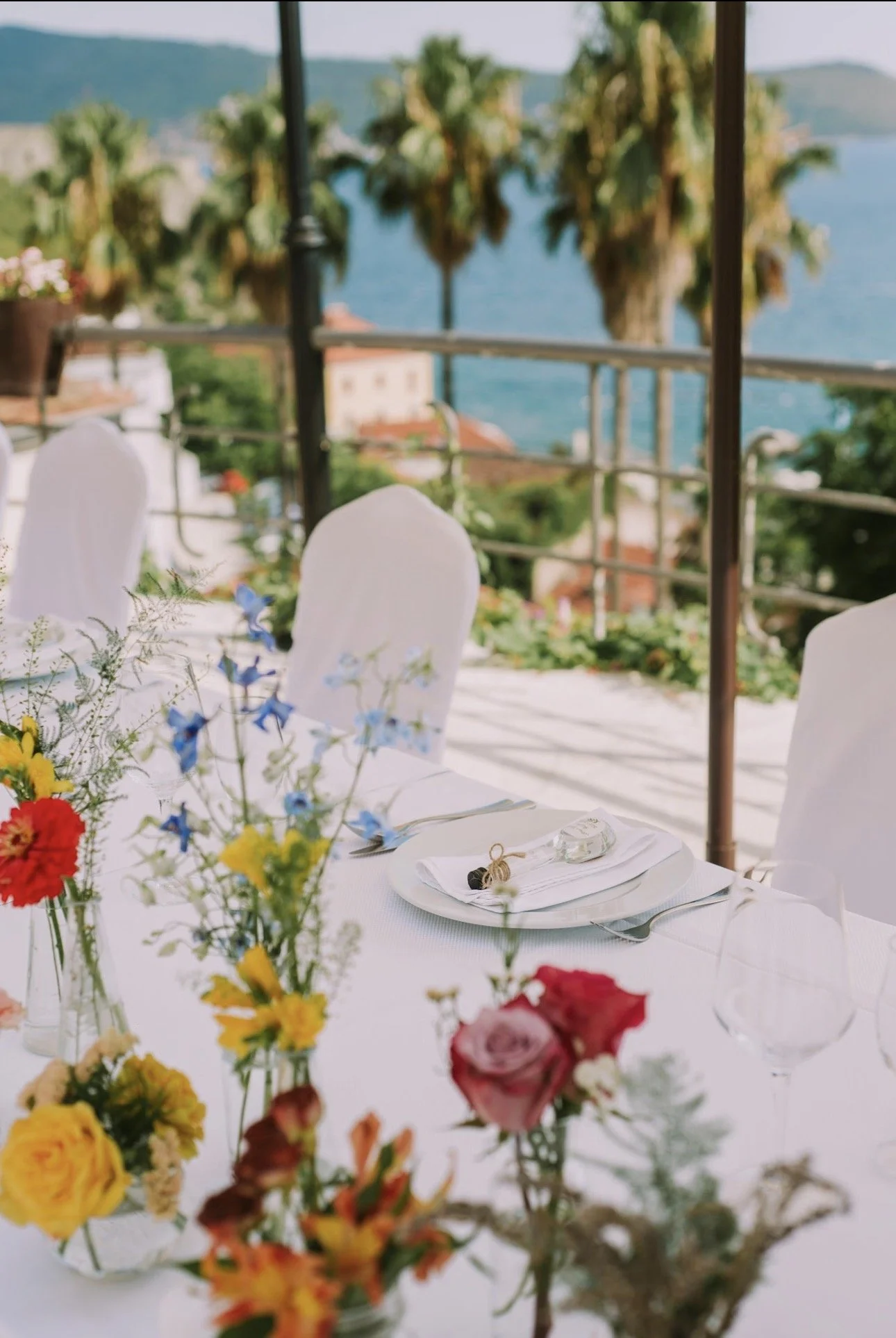 Decorated outdoor banquet table with colorful flowers, set for a special event, overlooking a tropical landscape with palm trees and water in the background.