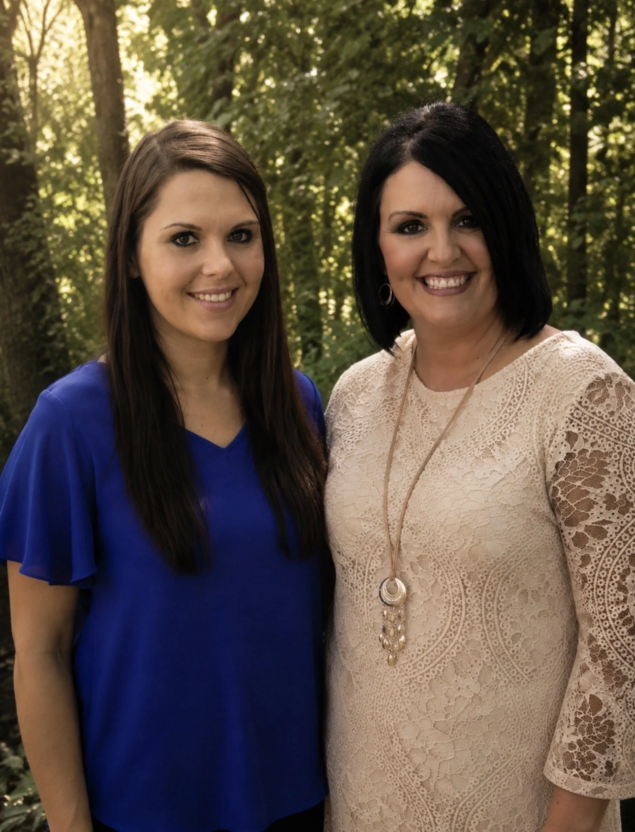 Two women smiling outdoors, surrounded by trees, one wearing a blue dress and the other in a beige lace dress with a long necklace.
