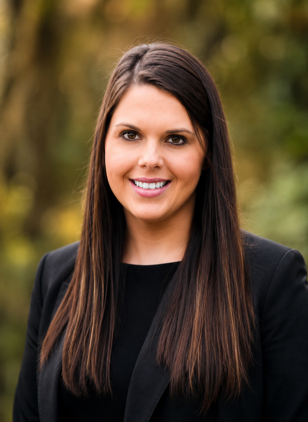 A woman with long dark brown hair, smiling, wearing a black blazer outdoors on a fall day.