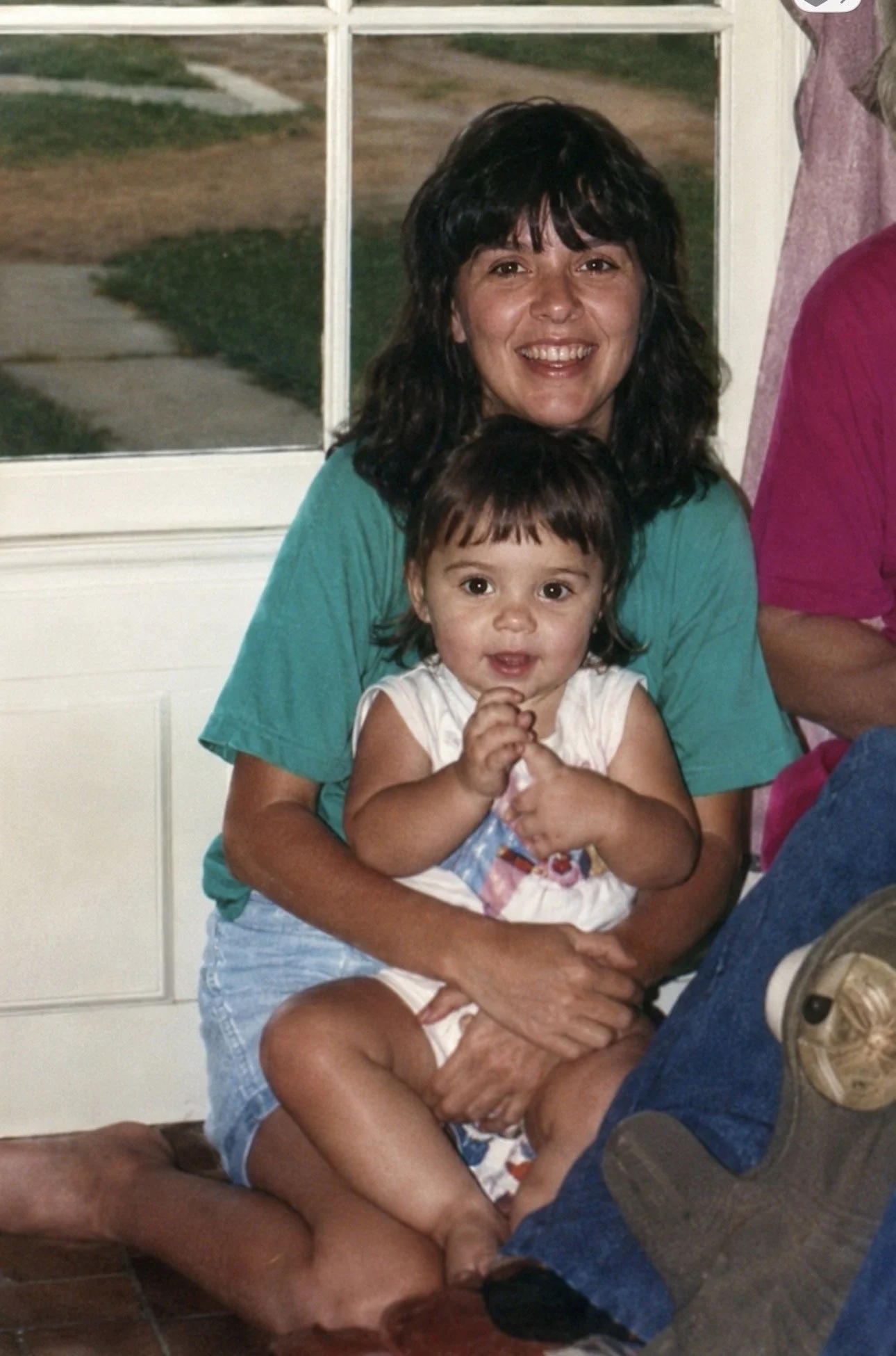 A woman with black curly hair smiling and holding a young girl with brown hair and bangs, who is clutching a toy or object, sitting on her lap inside a room with a large window showing an outdoor path and grass.